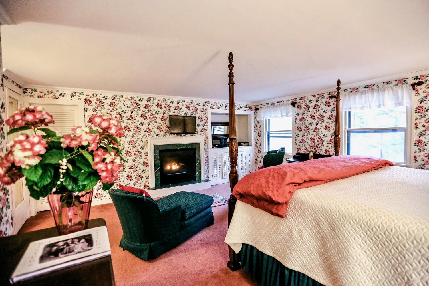 Cozy bedroom with floral wallpaper, a four-poster bed, a fireplace, and a seating area. Decor includes a vase of flowers.