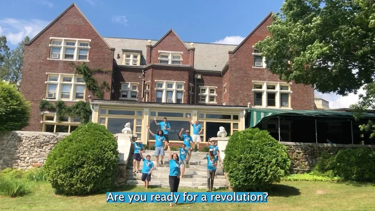 A group of people in blue shirts poses on the steps of a large brick building, with greenery around and a sunny sky above.