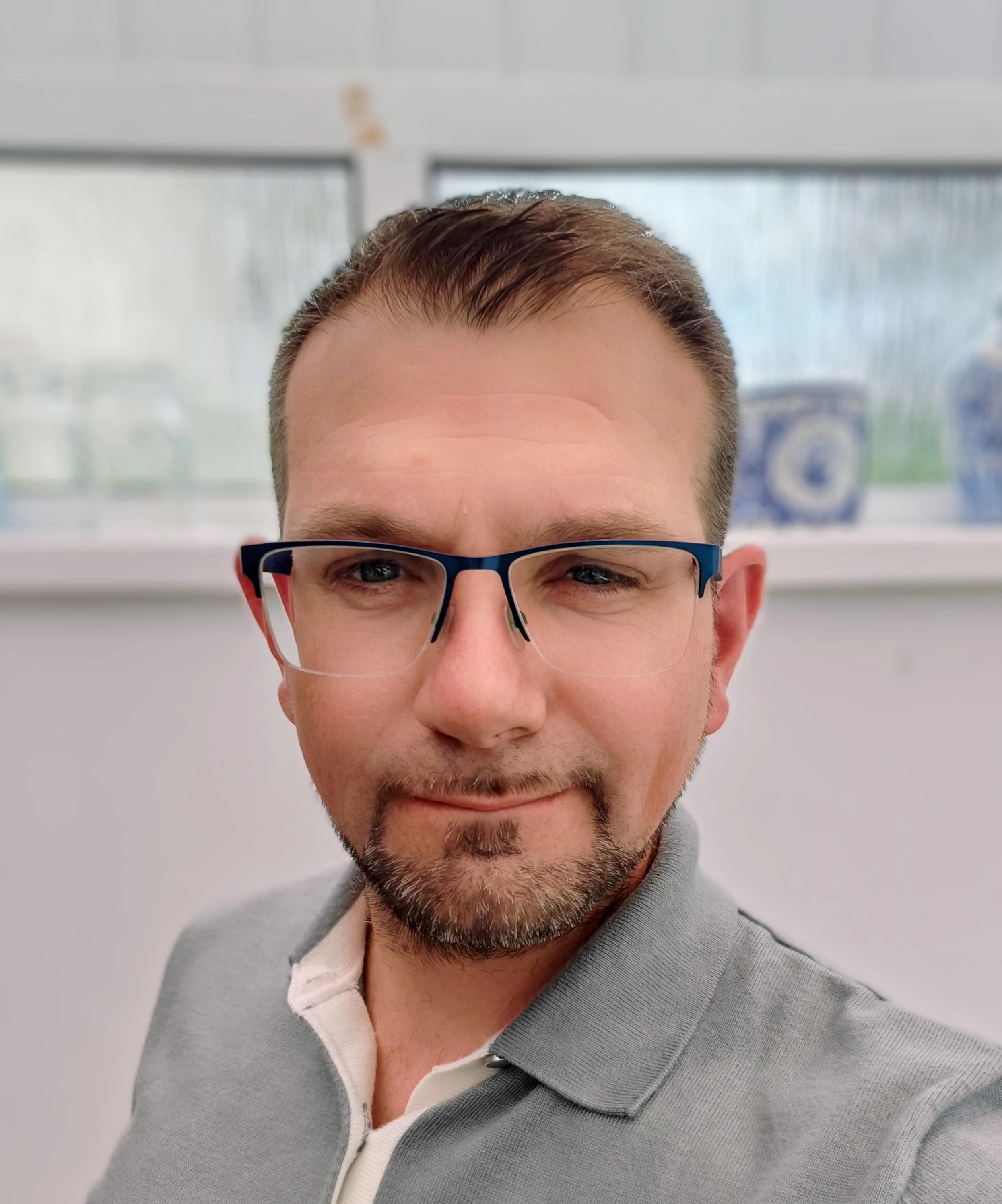 Close-up of a man with short brown hair, glasses, a neatly groomed beard, wearing a gray jacket and a white collared shirt, smiling, indoors with a blurred window in the background.