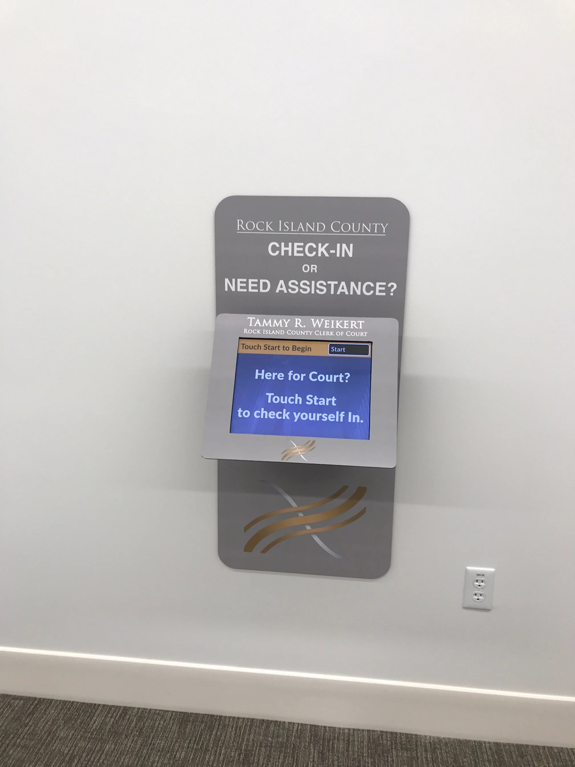 A sign on a wall that reads 'Rock Island County Check-in or Need Assistance? Tammy R. Weikert Rock Island County Clerk of Court.' The sign instructs users to touch start to check themselves in, with a digital screen displaying 'Here for Court? Touch Start to check yourself in.'