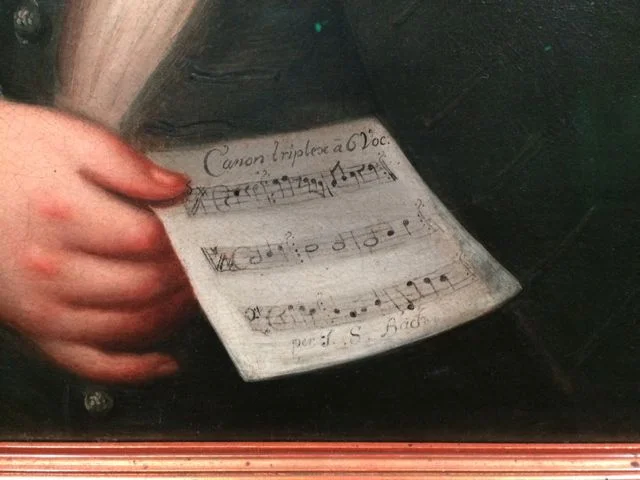 ...He is holding a code book for a canon in 6 voices; three are provided and the other three are to be solved.