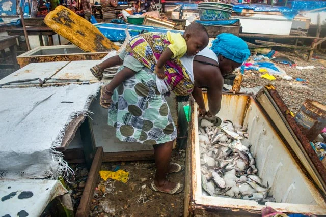 A woman picks some fish to sell them in the market while carrying her baby on the back.
