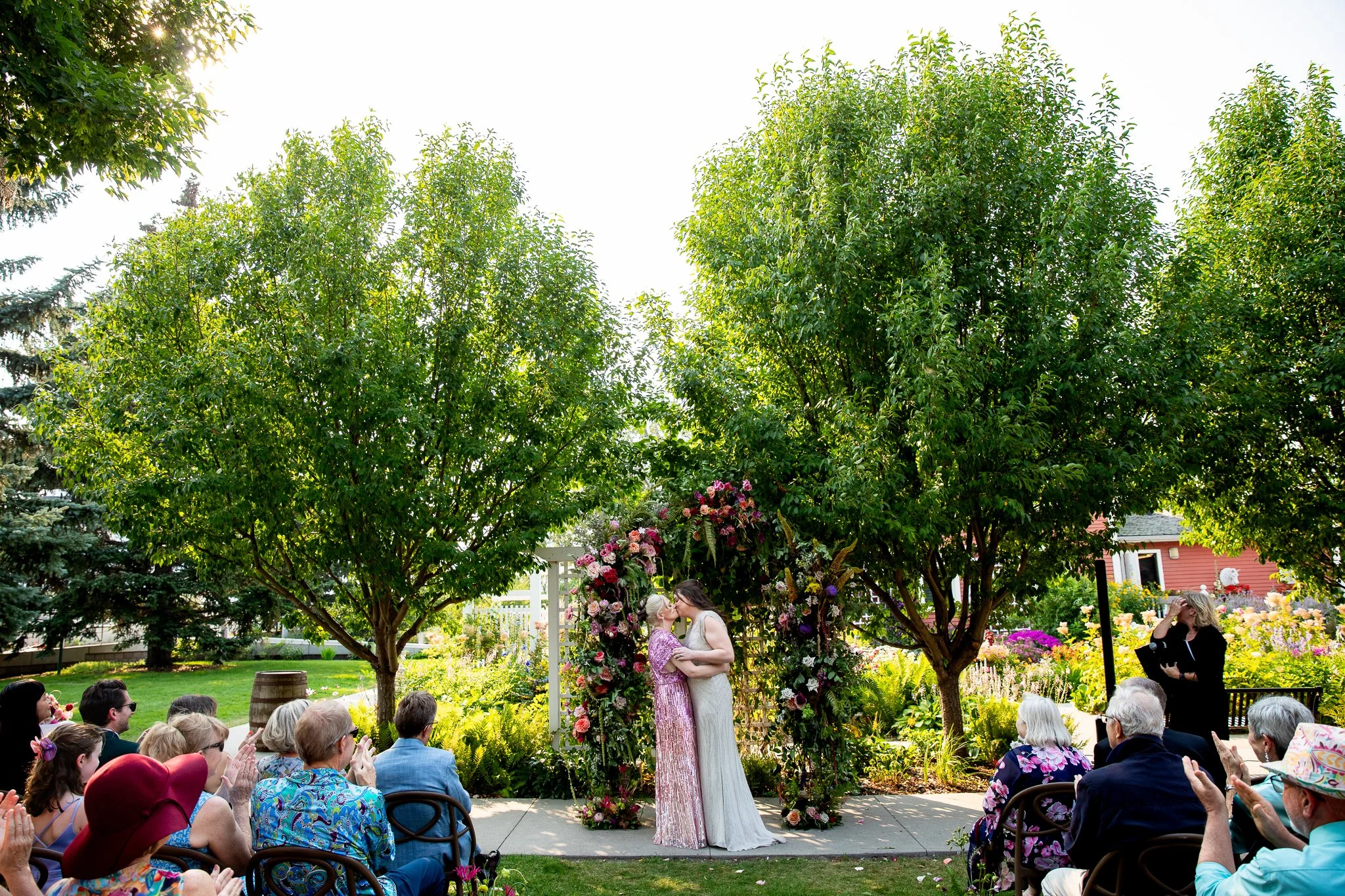 A same-sex couple shares their first kiss as newlyweds at The Deane House in Calgary, surrounded by cheering and applauding family and friends.