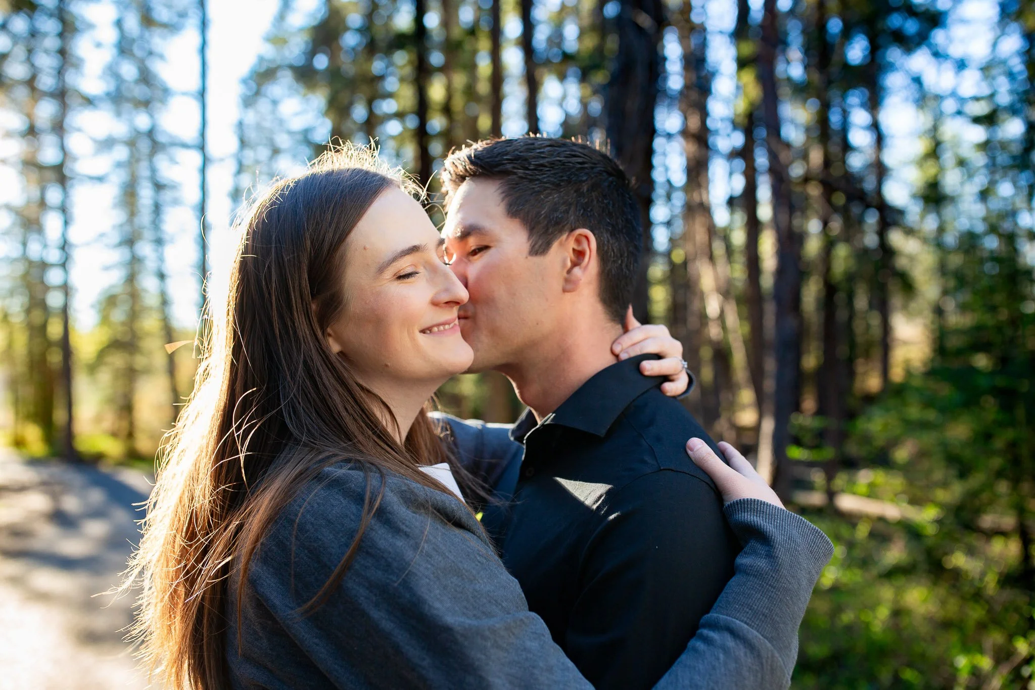 Engaged couple embracing among tall evergreens at Shannon Terrace in Fish Creek Park during sunset.