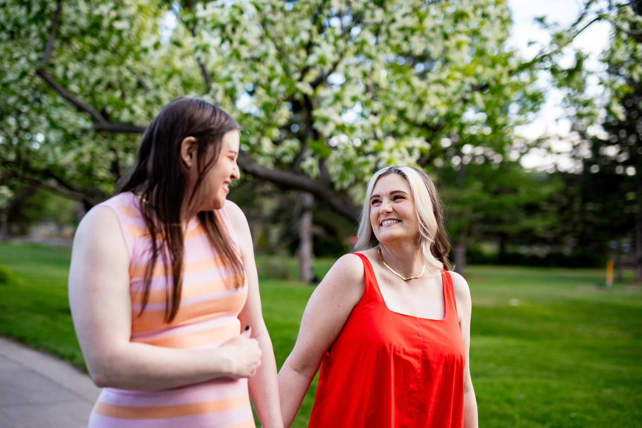 Dianna and Annika walk together beneath blooming lilac trees during their spring engagement session at Baker Park in Calgary.