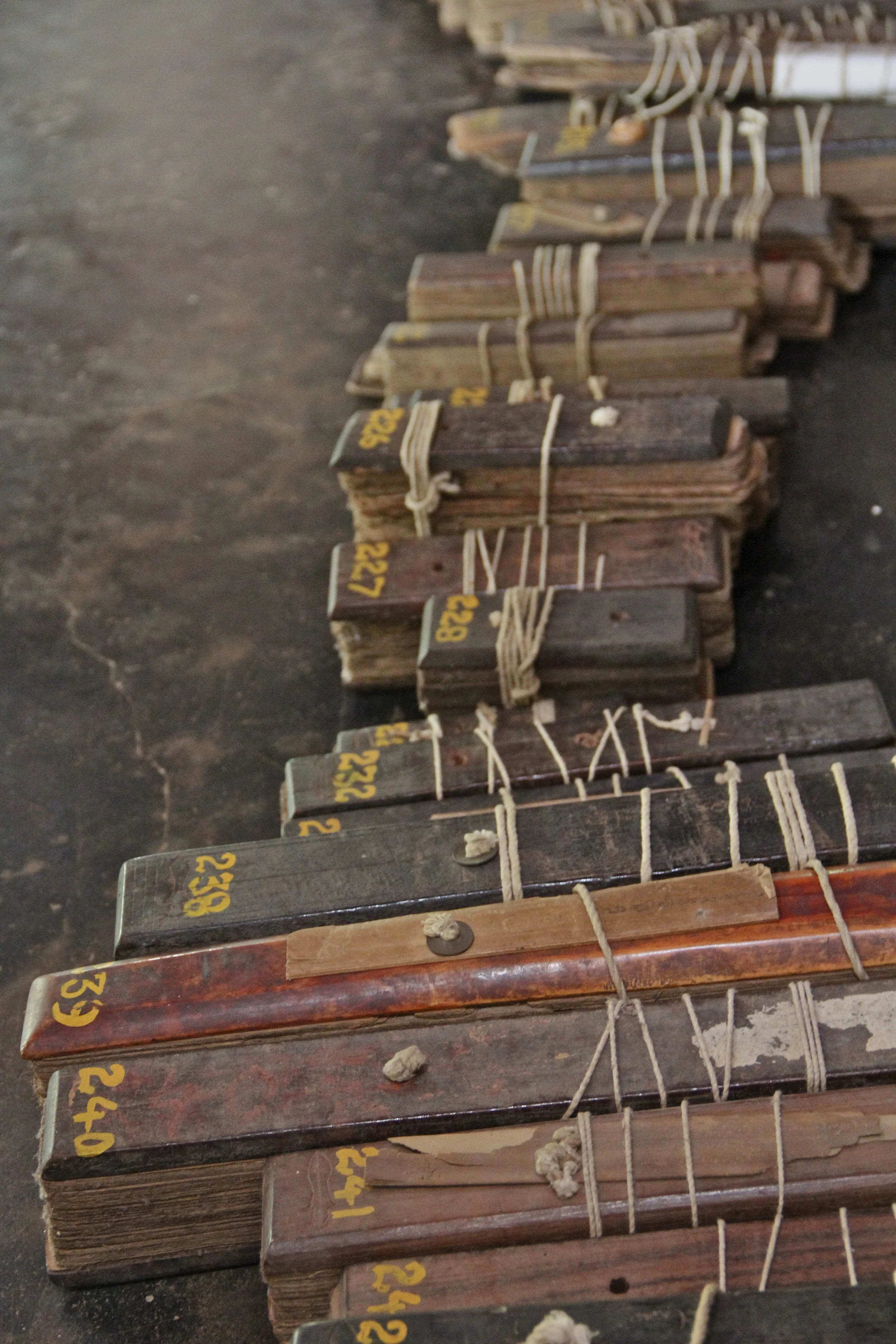 A small portion of a Kerala manuscript collection: stacks of palm-leaves, incised with text on both sides and stacked like cards, are clamped between wooden covers and tied with string threaded through holes in the middle. Each "book" has been numbered in yellow by modern researchers...