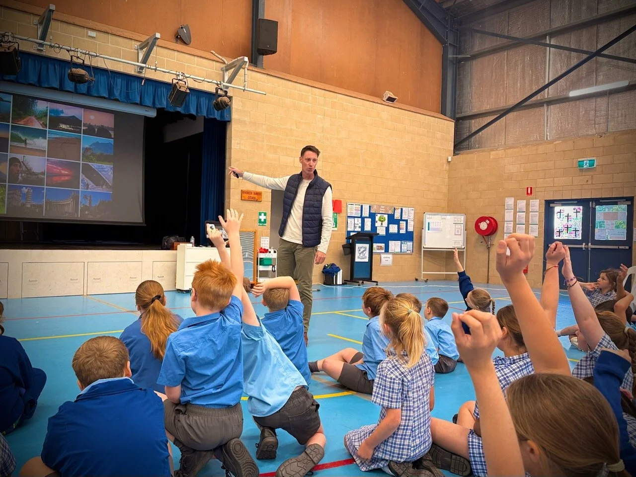 Sam Clear instructs a group of students seated on the gymnasium floor, with several students raising their hands. The students are wearing school uniforms, and Sam is pointing at a projection screen displaying landscape images from his walk.