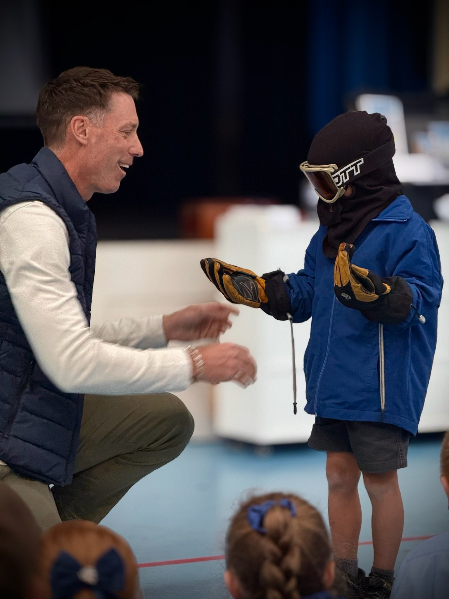 Sam Clear kneeling and smiling while talking to a child wearing ski goggles and winter clothing. The child is gesturing as they converse in an indoor setting, with other children watching.
