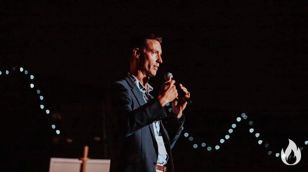 Sam Clear in a suit holding a microphone on stage with decorative lights in the background, speaking at a Catholic conference.