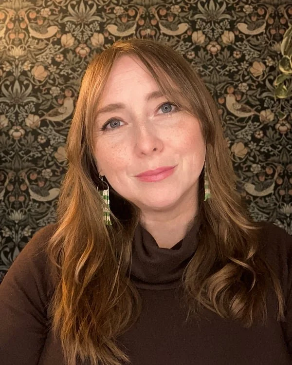 Portrait of Nicole Lange with long reddish brown hair, wearing a brown turtleneck shirt and dangly earrings, in front of a wallpaper with plants and birds.