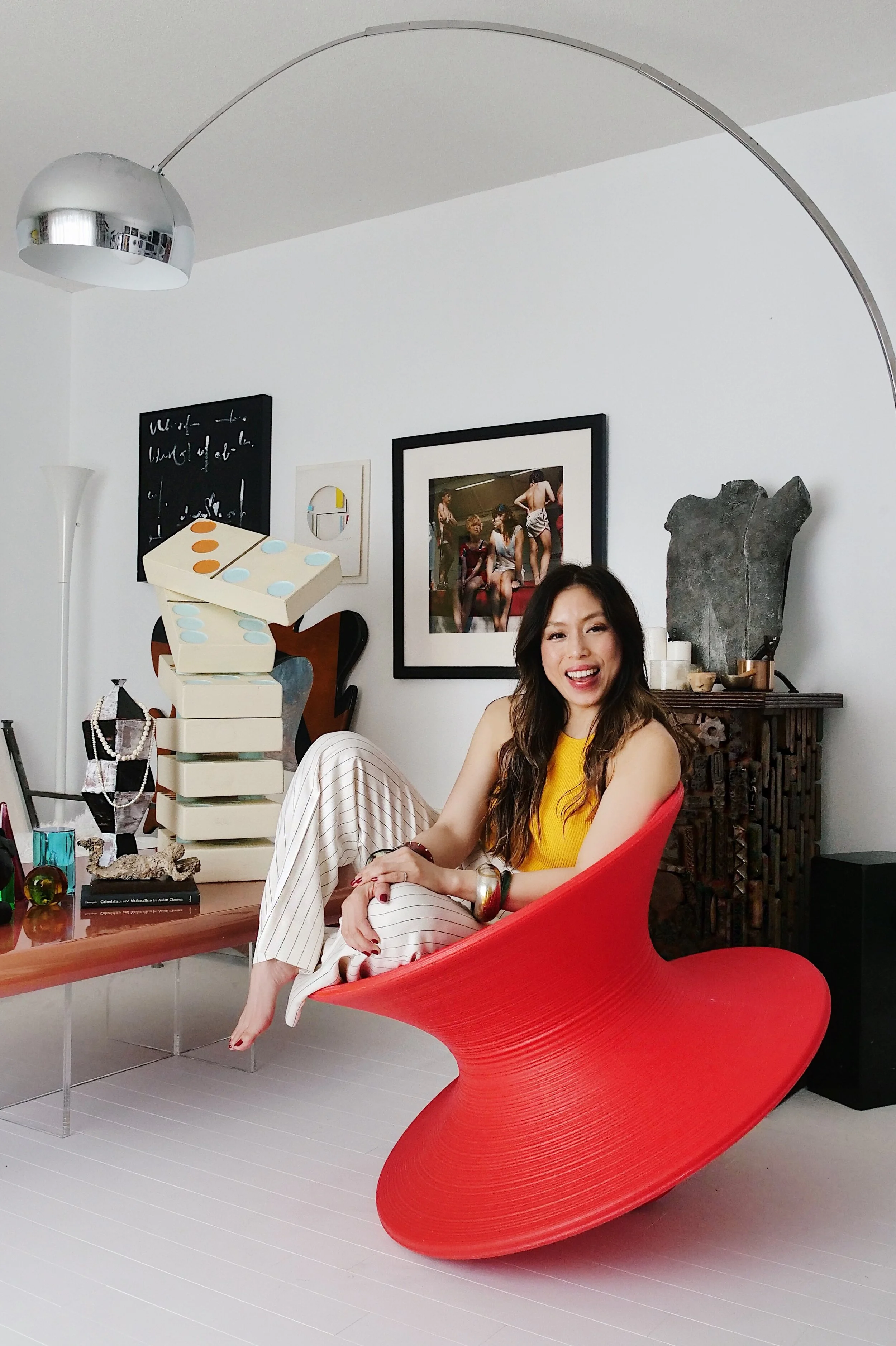 A woman with long dark hair smiling and sitting on a modern red chair in a contemporary living room with art and decorative items.