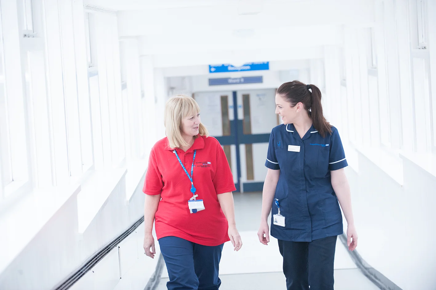 Two nurses walking down hospital corridor smiling at each other