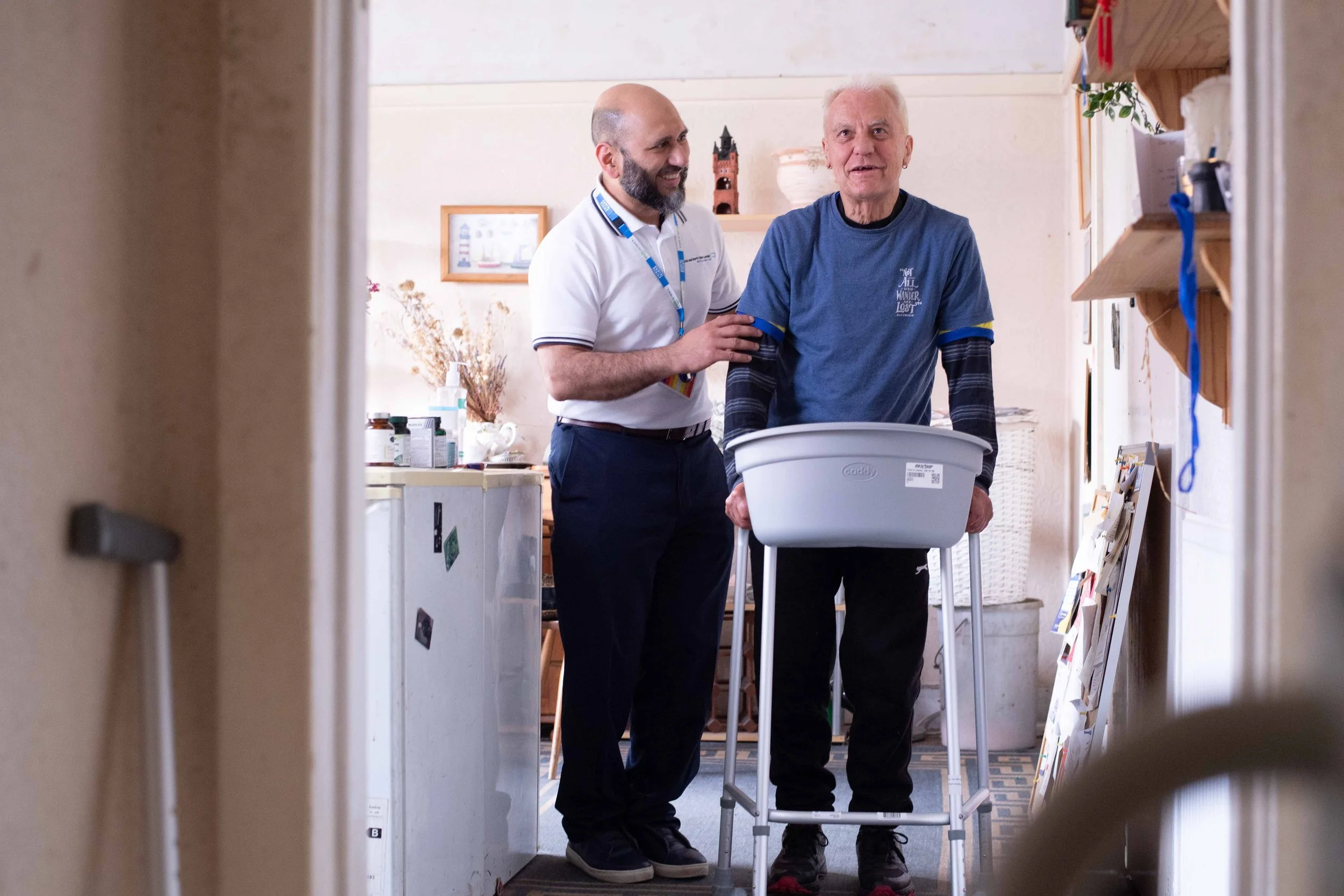 A male NHS Physiotherapist helps a man, in his own home, walk with a walking frame. It's a positive scene, with both men smiling, Healthcare Photography