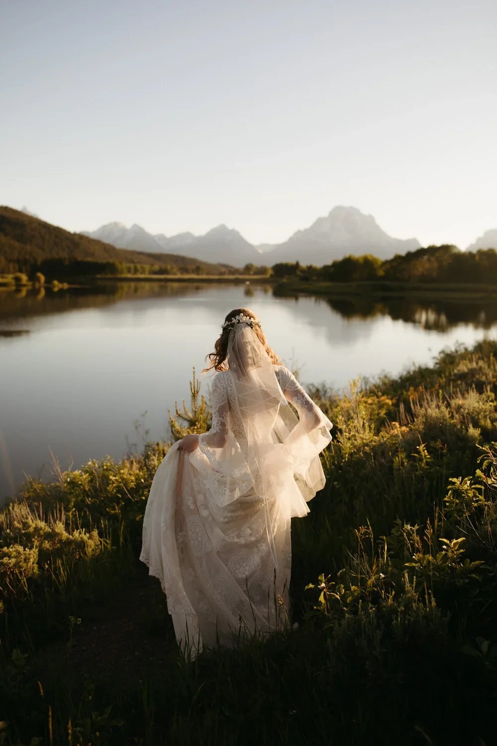 Bride walking toward a calm lake at sunrise with soft light and the Tetons reflected in the water.