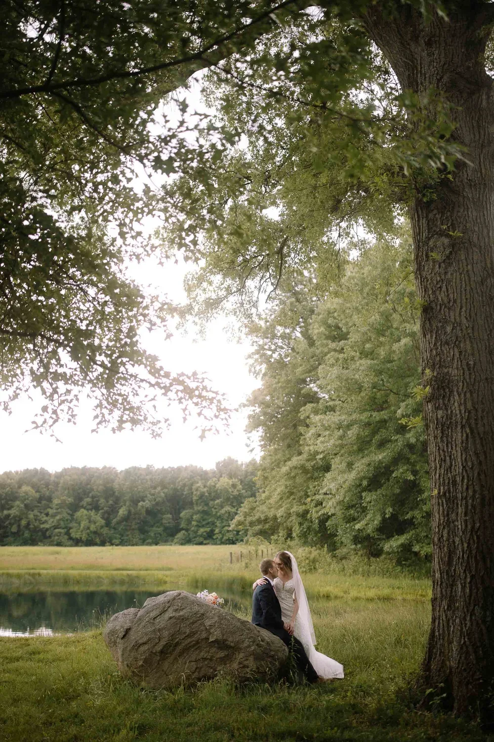 Bride and groom standing beside a large rock by a quiet lake during wedding portraits.