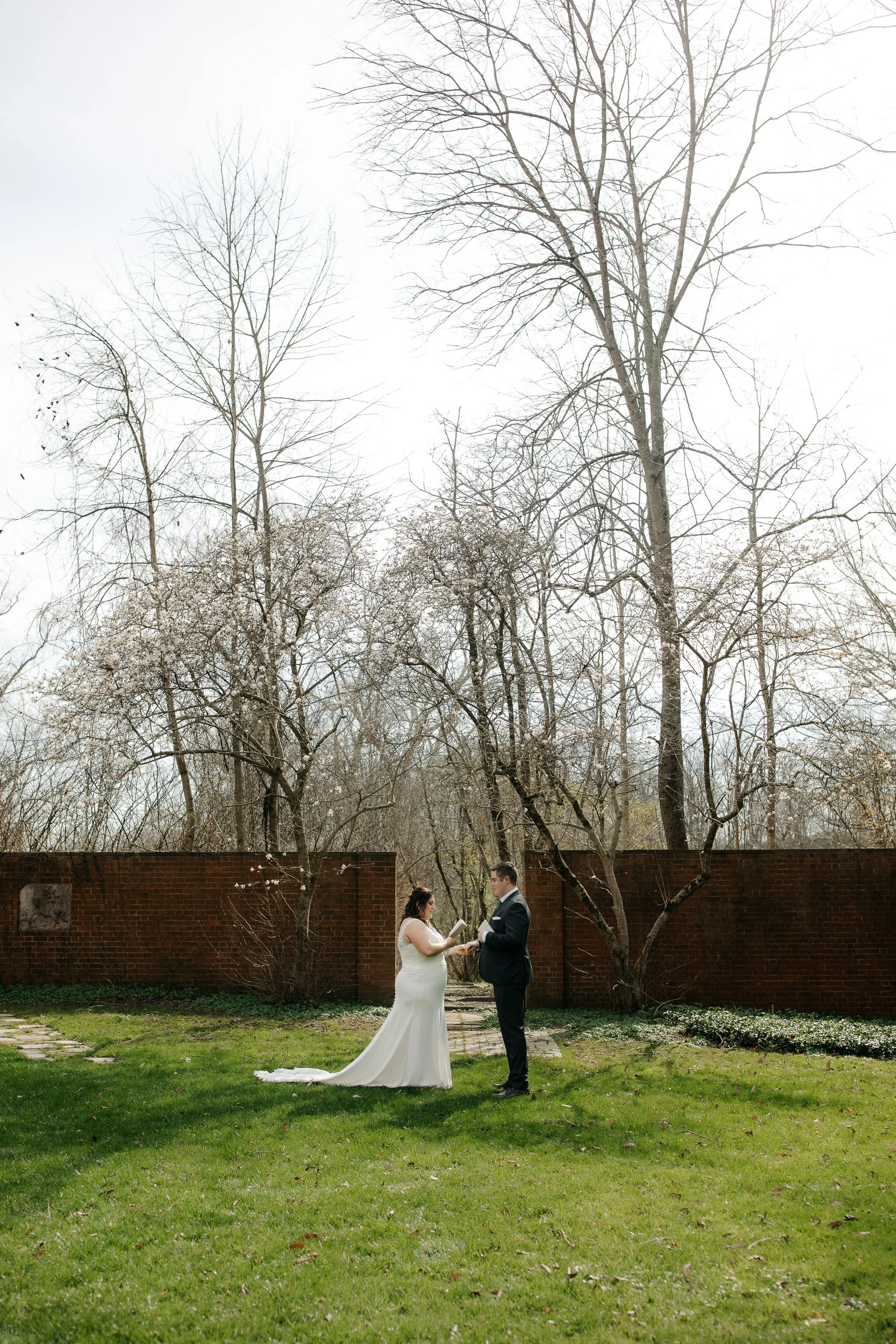 Wide view of couple exchanging vows in garden with brick wall backdrop at Peterloon Estate
