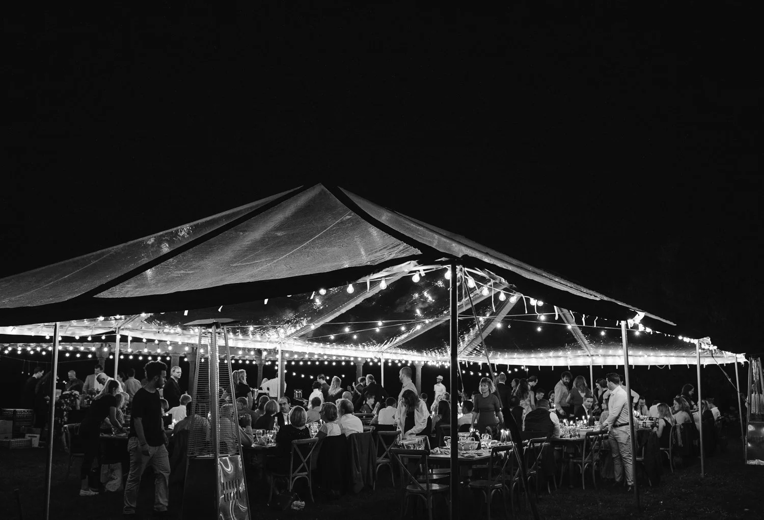 Wide black and white view of outdoor wedding reception under tent showing guests interacting and layered evening atmosphere