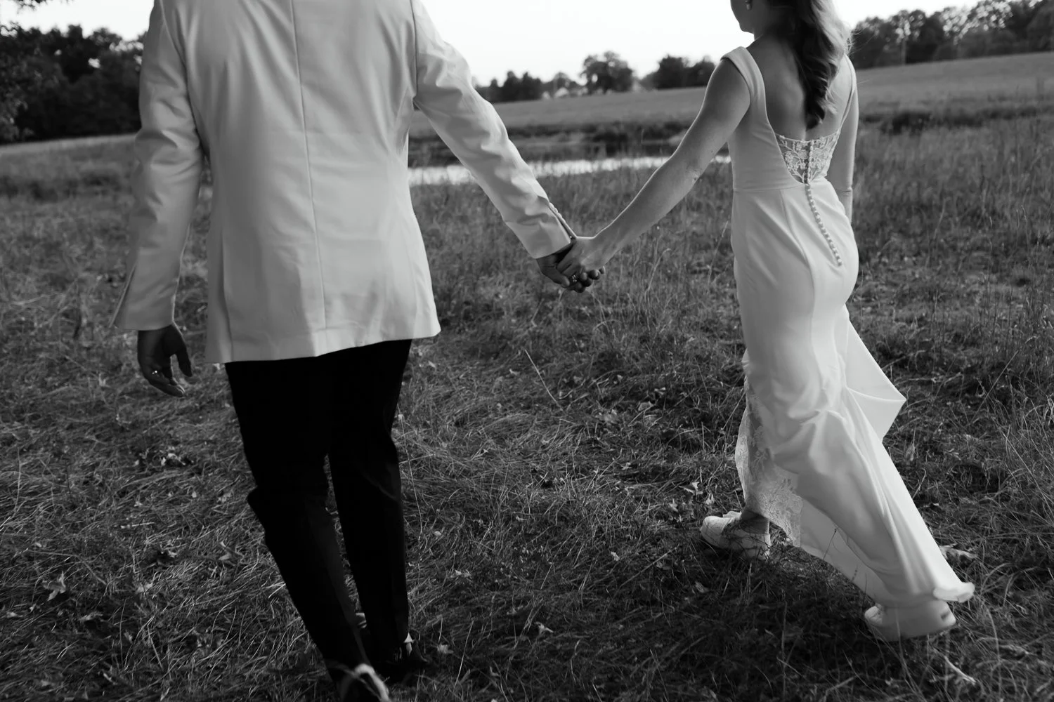 Black and white candid portrait of bride and groom walking hand in hand through an open field