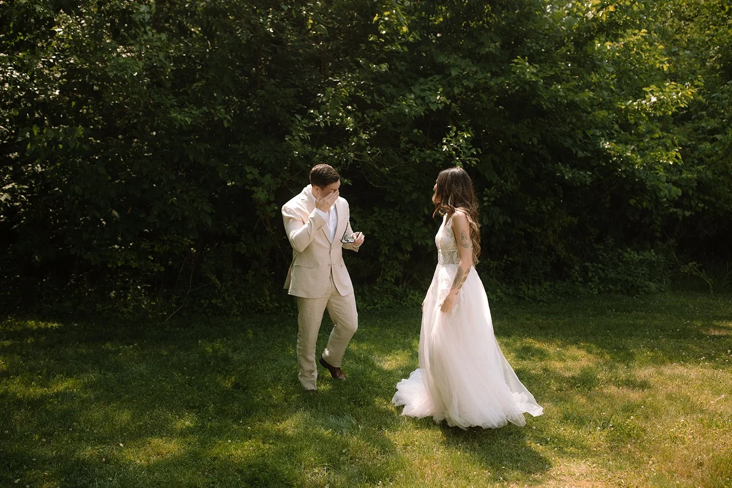 Groom wiping away tears during a private first look as the bride approaches him outdoors.