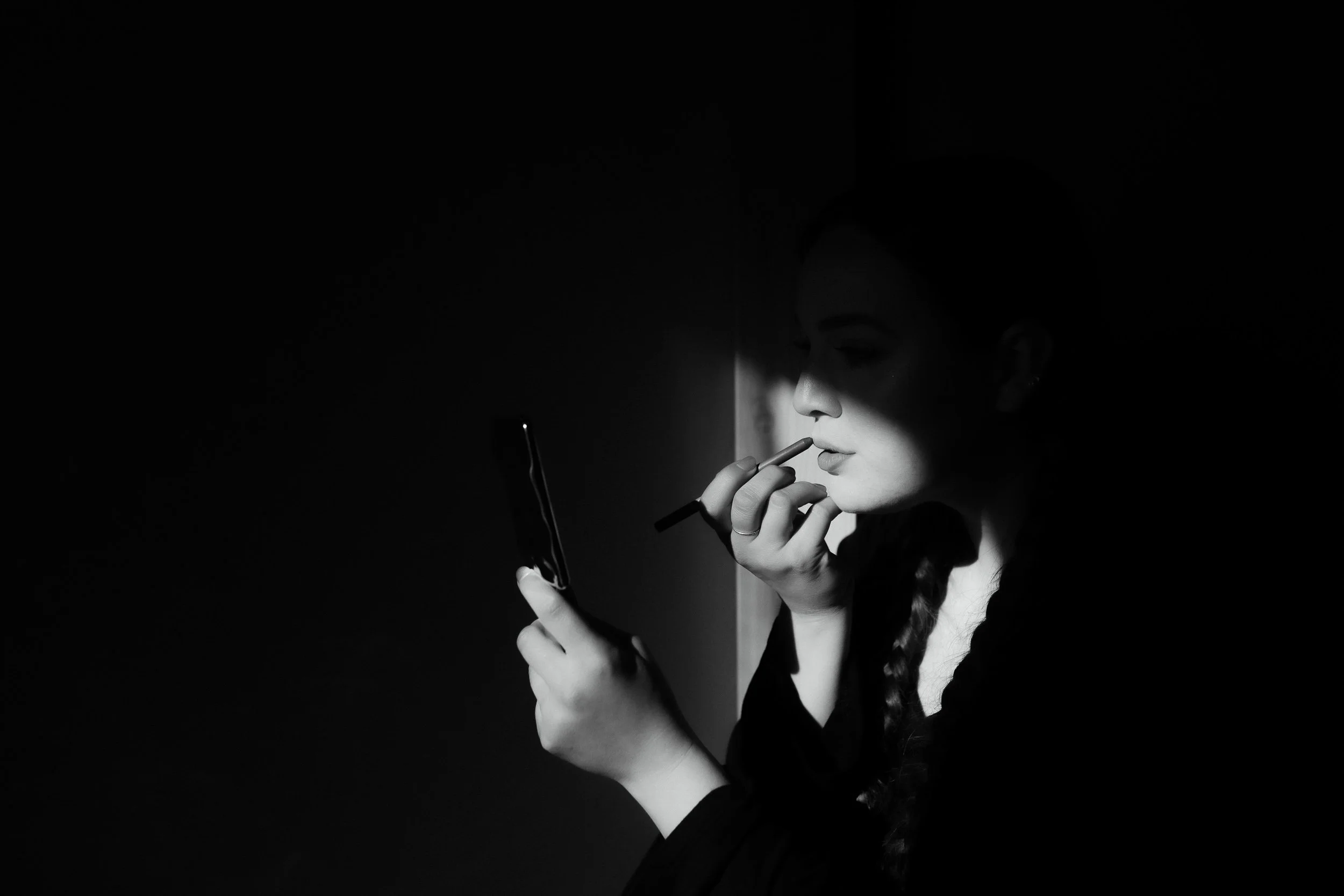 Black and white portrait of bride applying lipstick in dramatic window light before the ceremony.