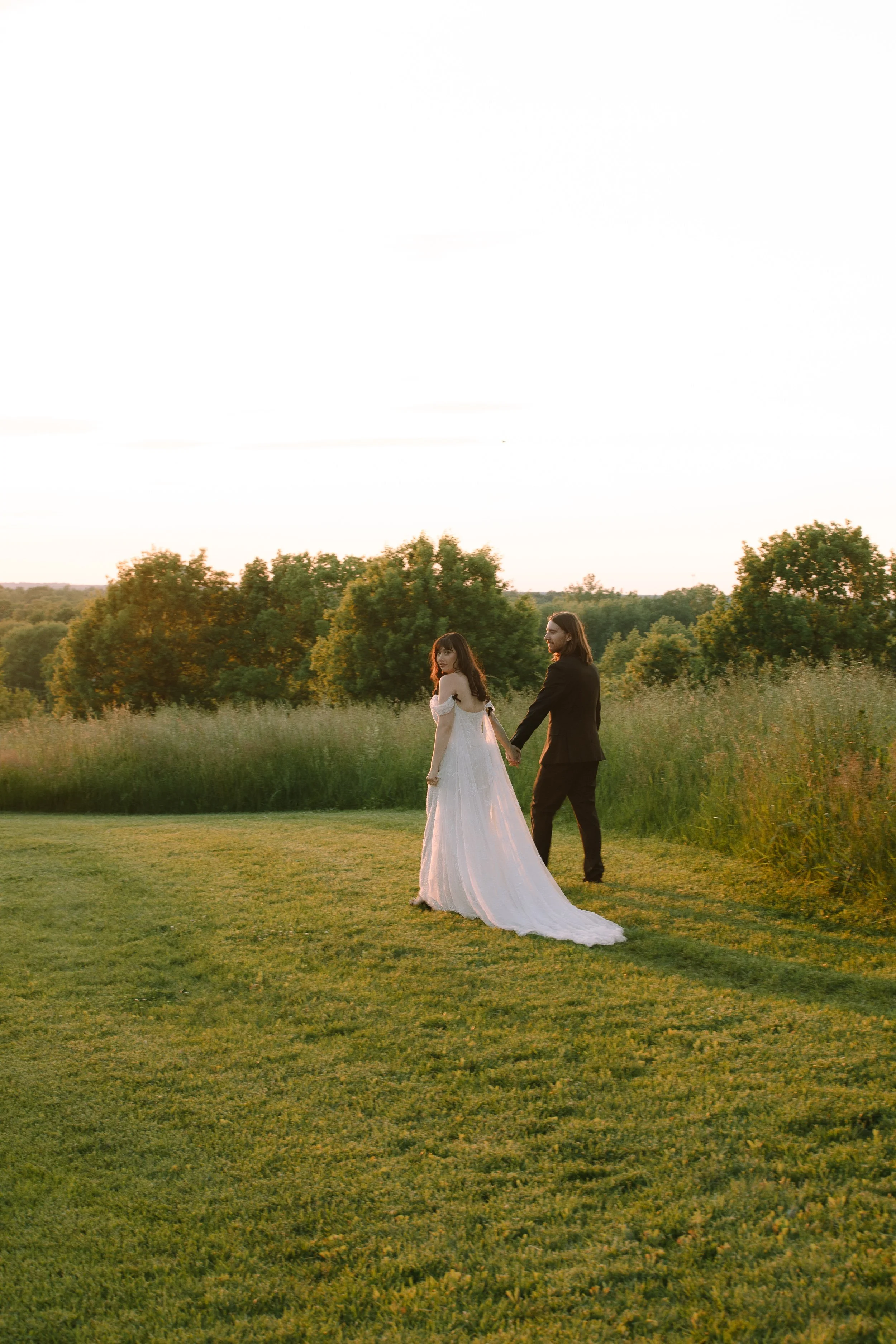 Couple walking hand in hand through tall grass at sunset during golden hour wedding portraits.