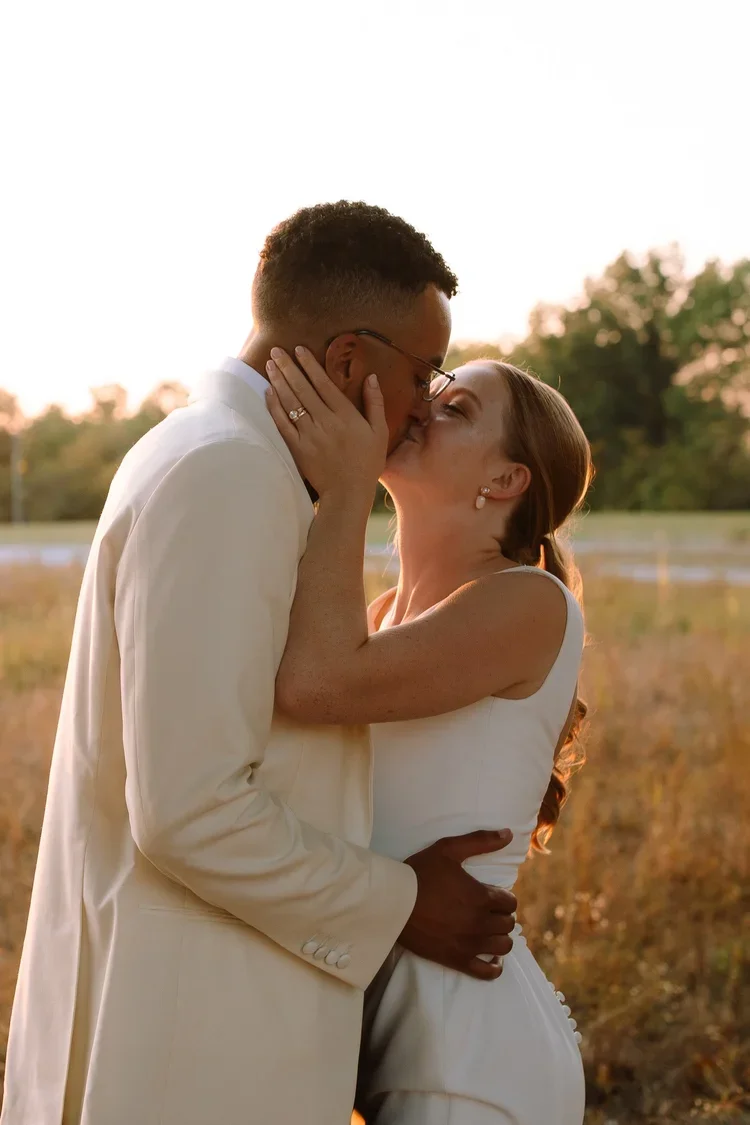 Bride and groom kissing in warm golden light during sunset wedding portraits in an open field.