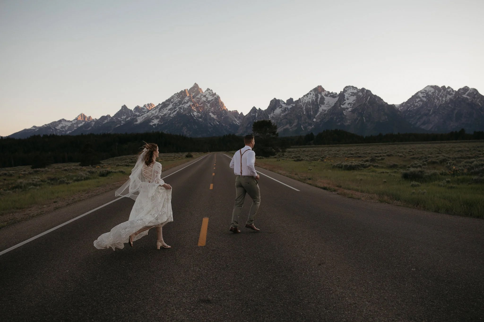Couple crossing an open road at sunset with the Teton mountain range in the background during their elopement