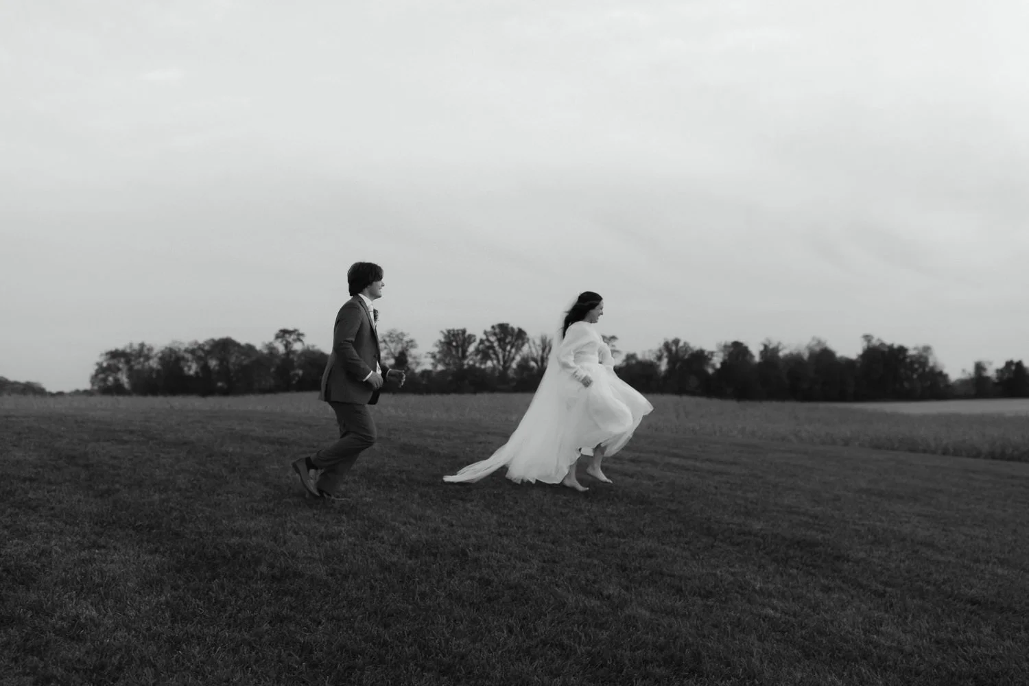 Candid black and white photo of a bride and groom walking through a grassy field at Ivory Meadows during their autumn wedding.