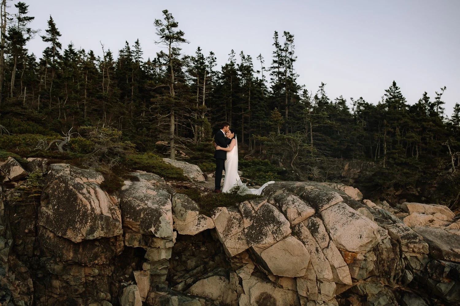Couple standing together on coastal cliffs surrounded by forest and stone during an Acadia elopement
