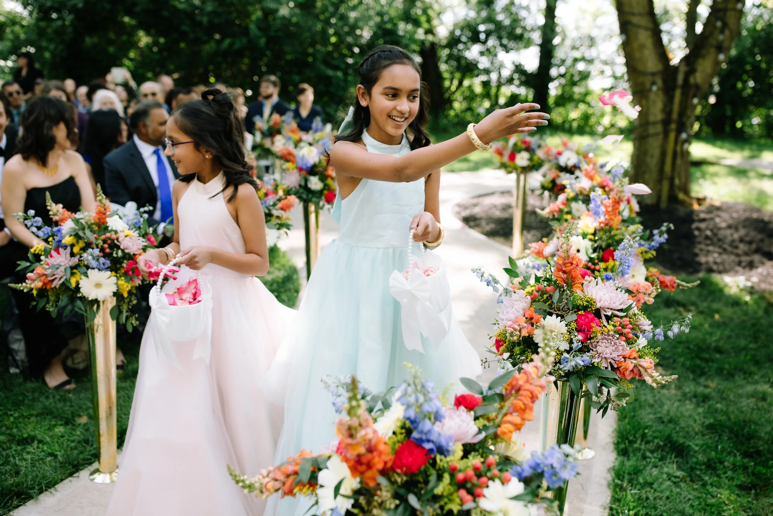Flower girls tossing petals down a colorful floral-lined aisle during an outdoor wedding ceremony.