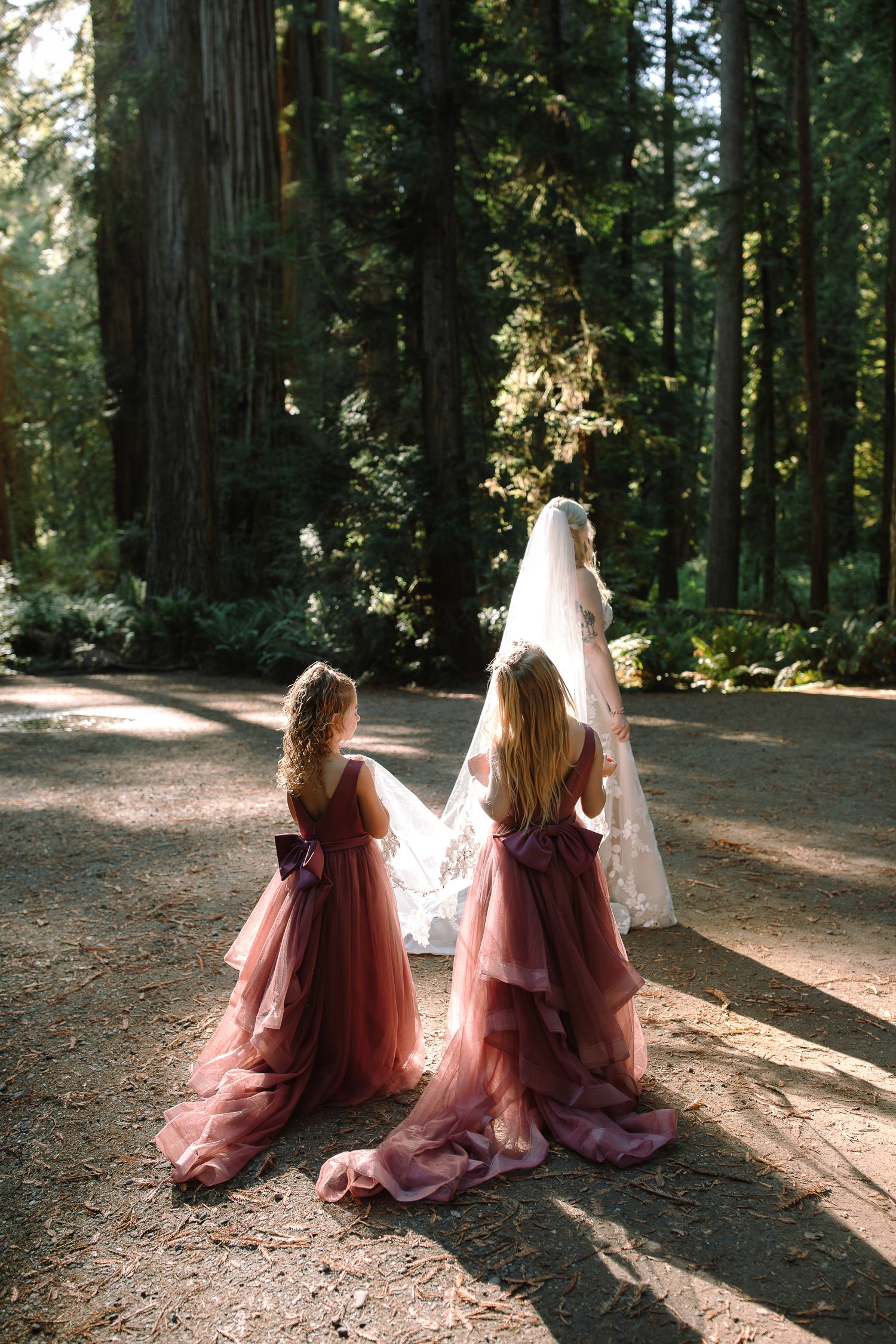Flower girls watching bride walk through redwood forest aisle