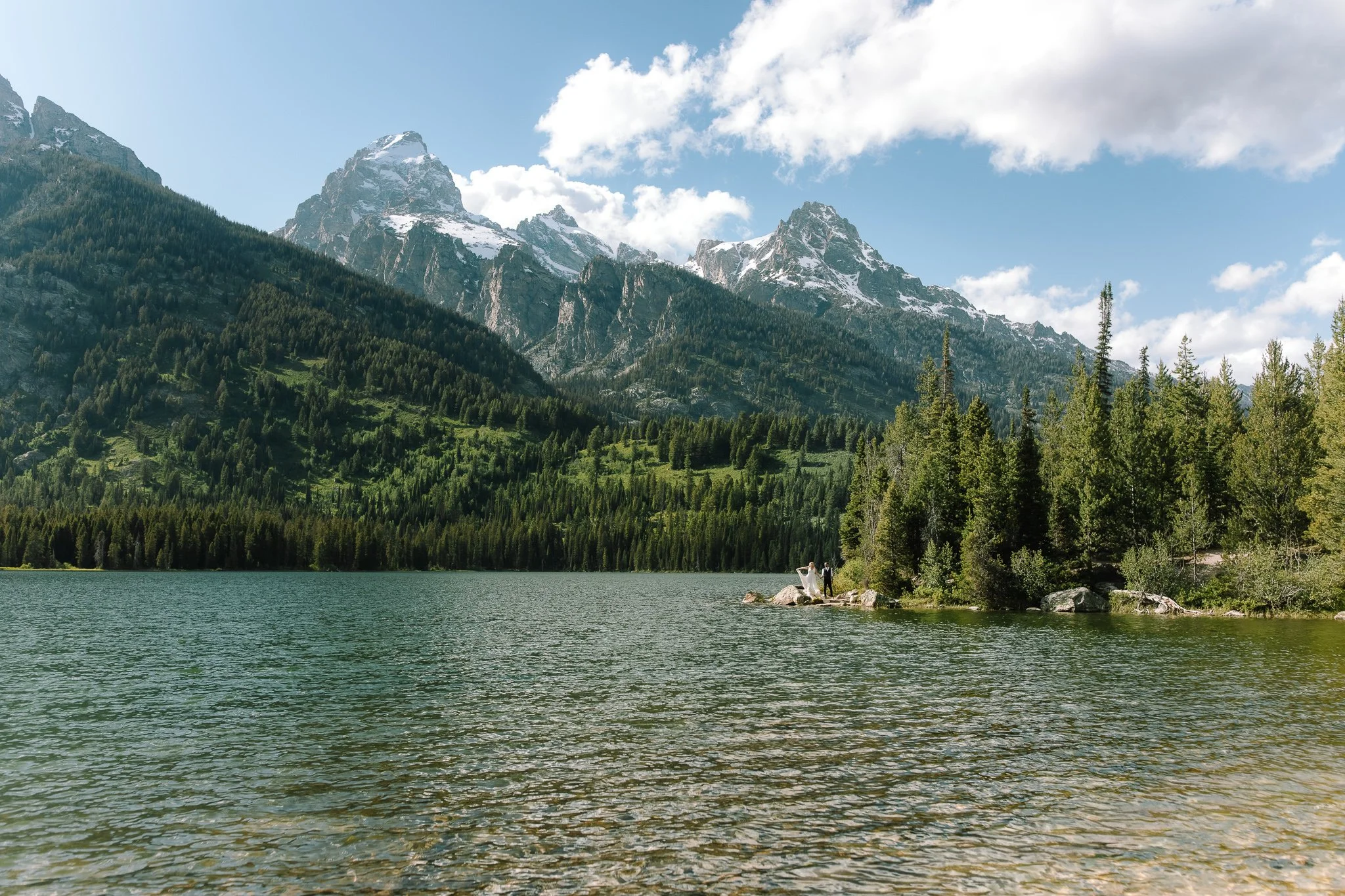 Wide view of a couple standing along a lakeshore in Grand Teton National Park surrounded by mountains