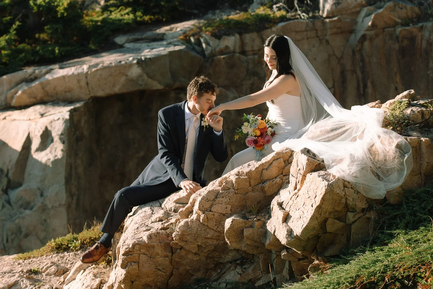 Intimate elopement portraits of a bride and groom seated together on rocky cliffs along the Acadia coastline