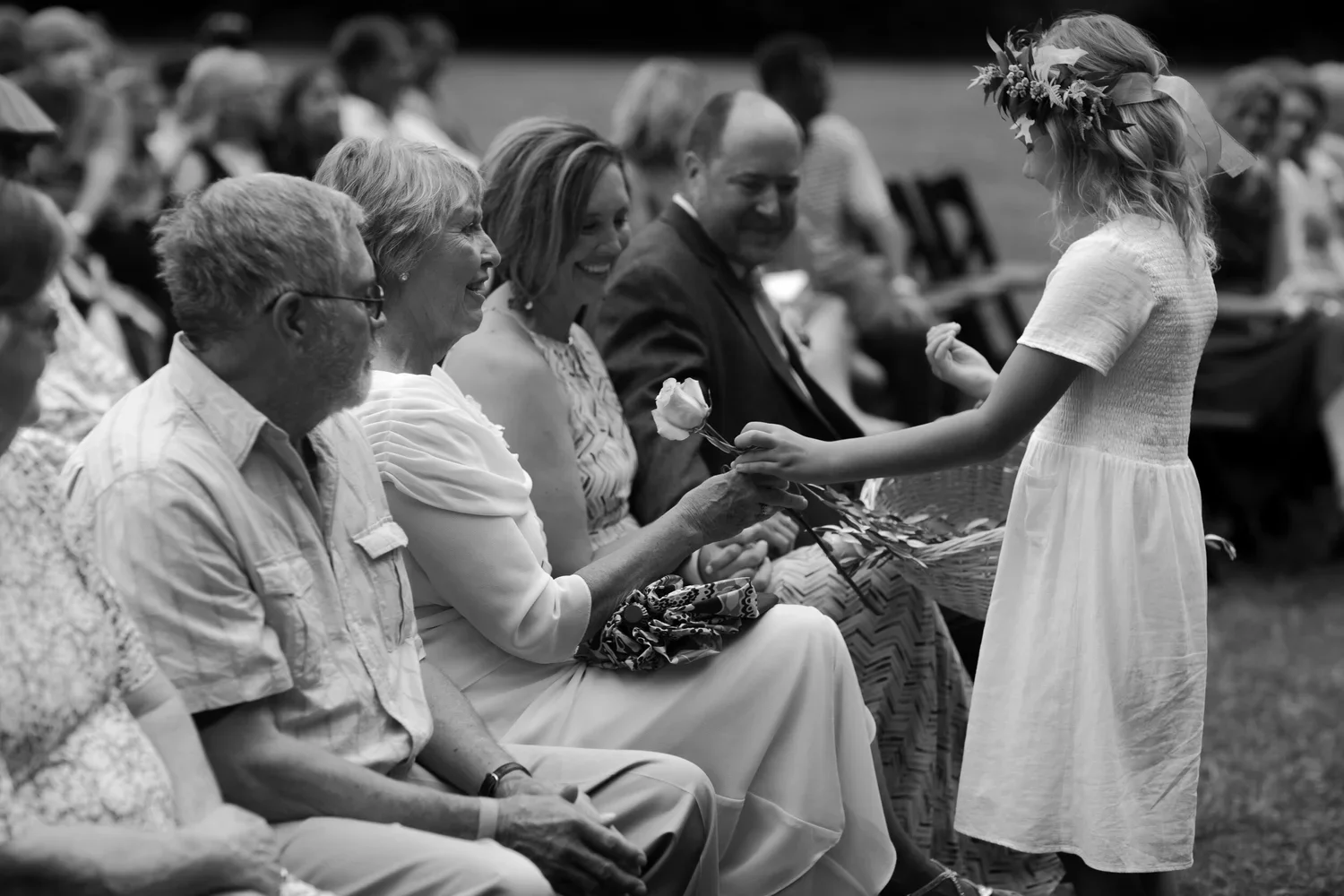 Young flower girl handing rose to seated family members during emotional outdoor wedding ceremony