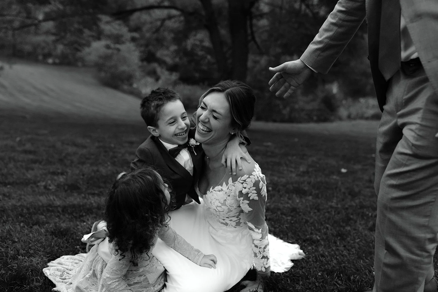 Bride laughing while sitting on the grass with two young children during family portraits.