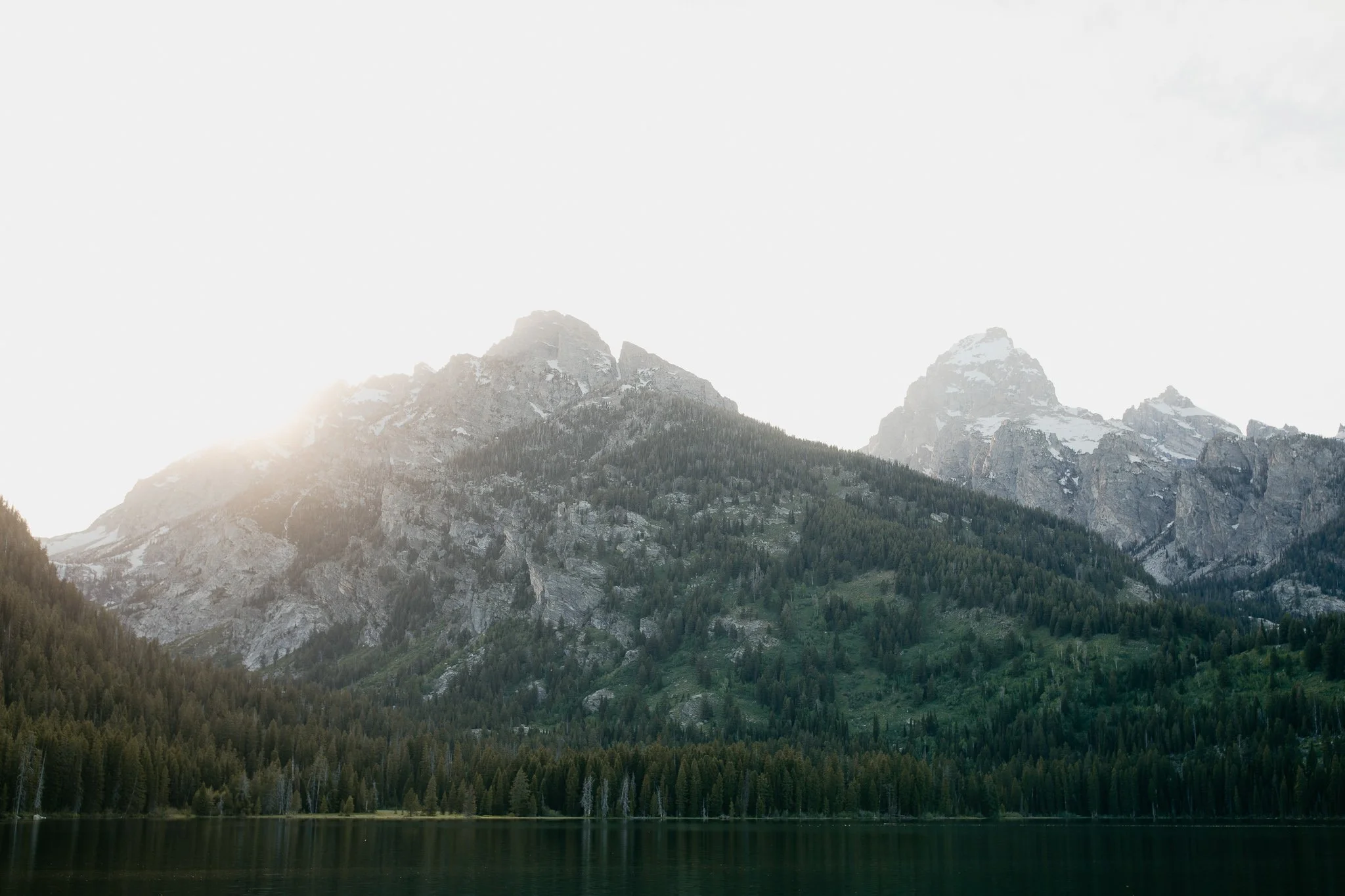 Soft light falling across the Teton mountain range with evergreen forest and lake in the foreground