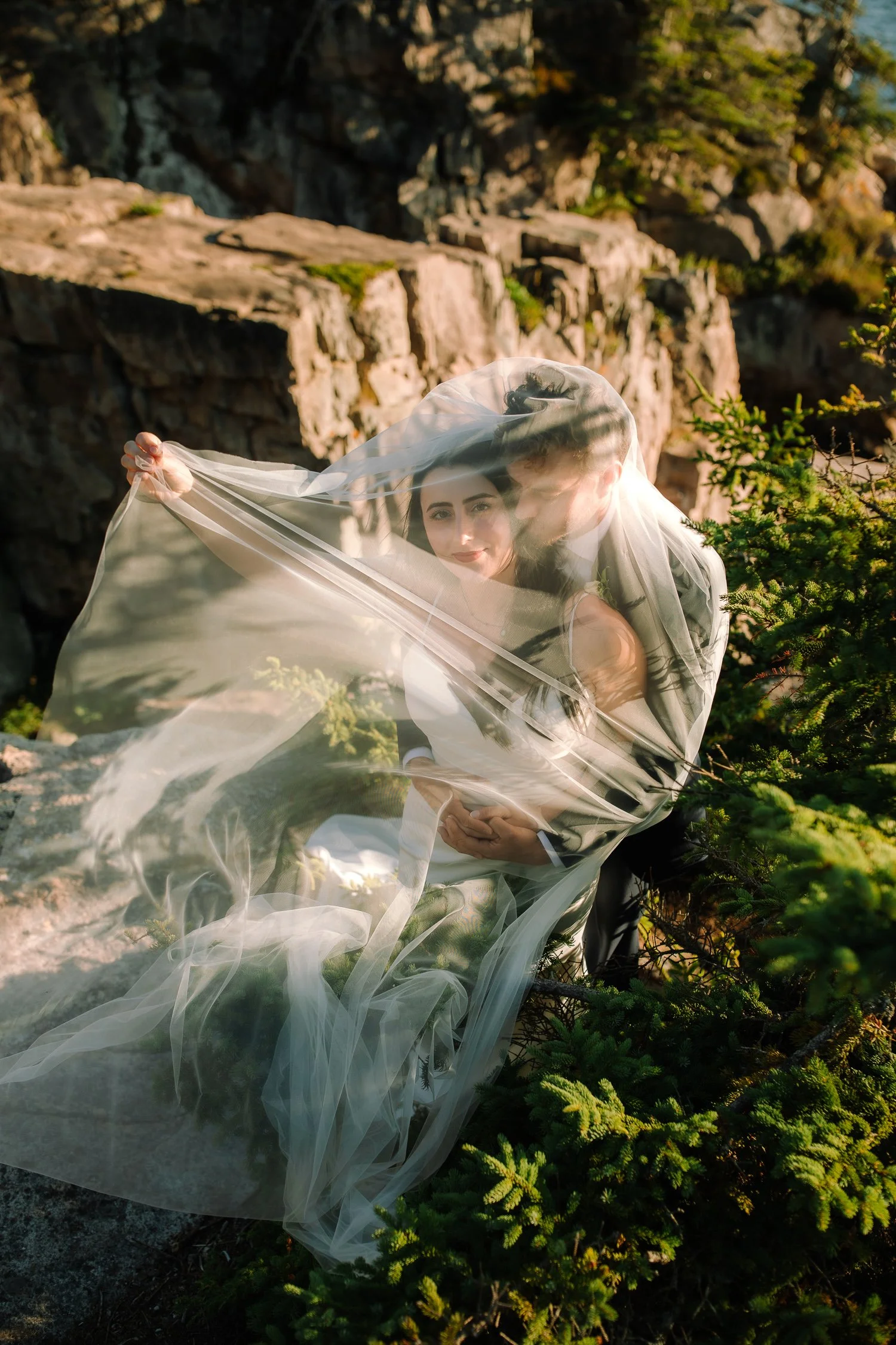 Artistic elopement portrait of a bride and groom wrapped in a veil among rocky cliffs and evergreens in Acadia National Park