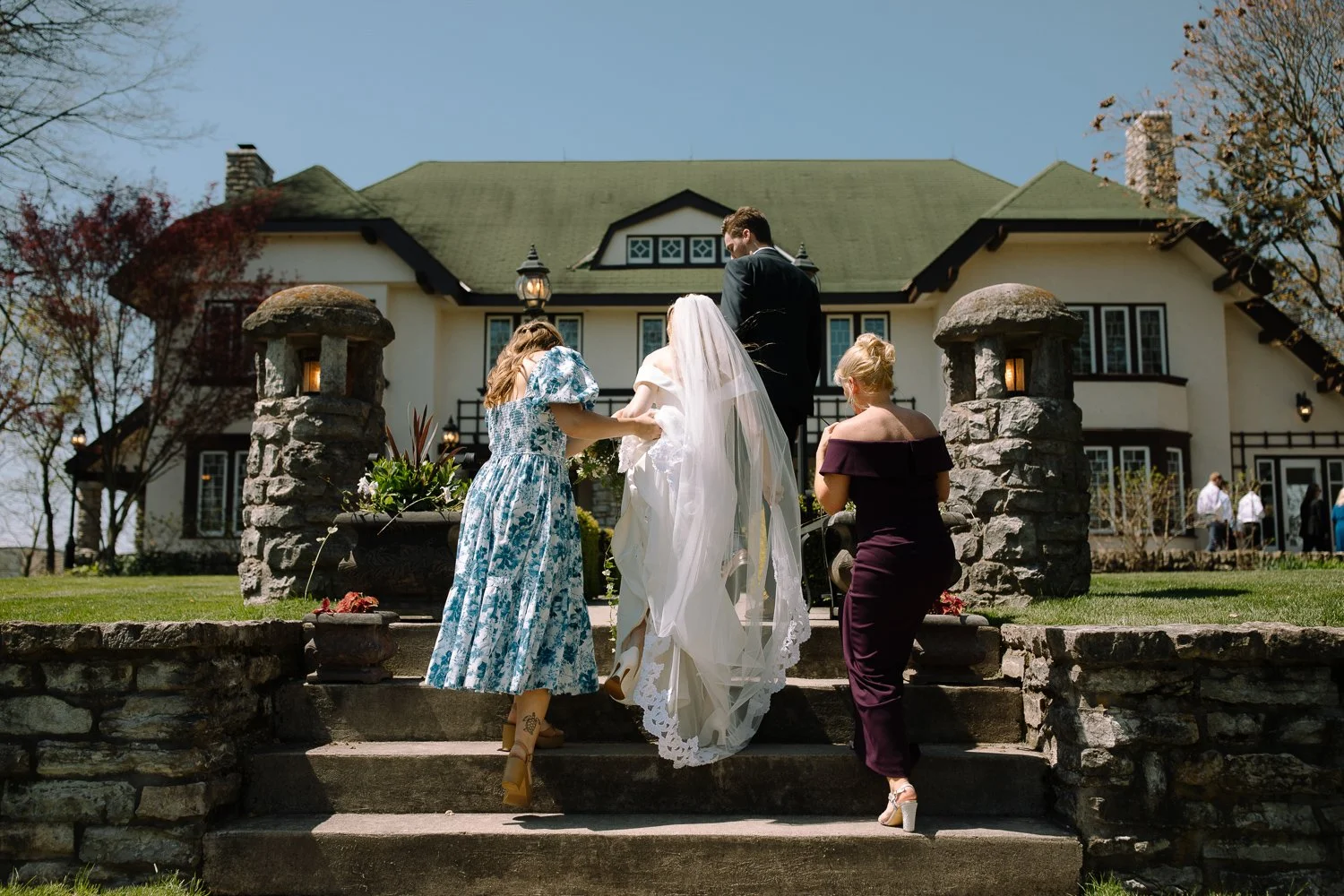 Bride walking up stone steps toward the ceremony with bridesmaids helping with her dress