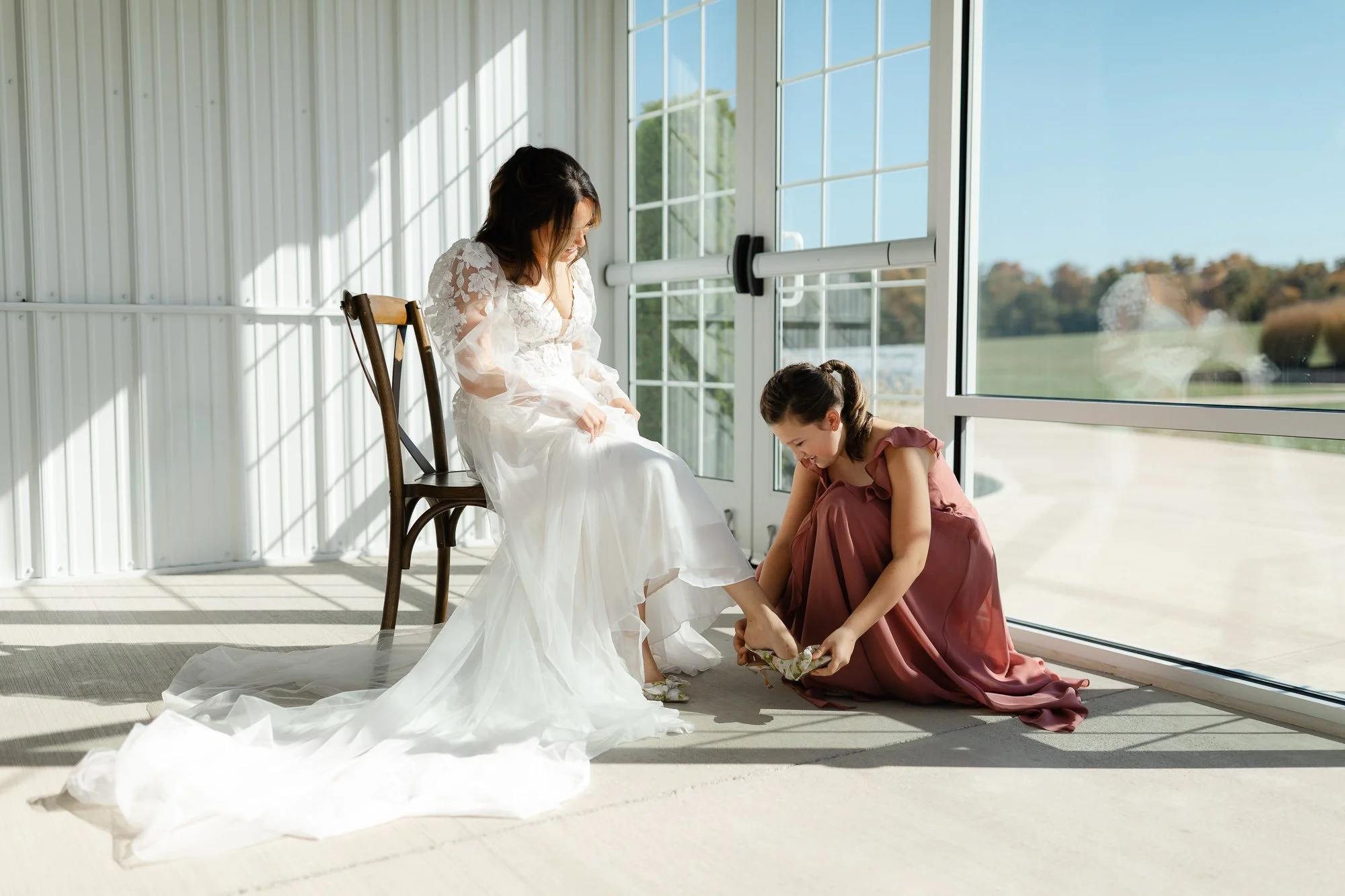 Bride seated by large windows while a bridesmaid helps her put on her wedding shoes.