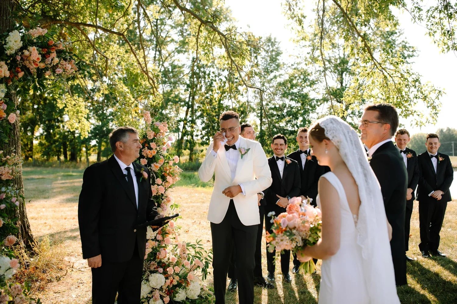 Groom reacting emotionally during an outdoor wedding ceremony beneath a peach and blush floral arch.