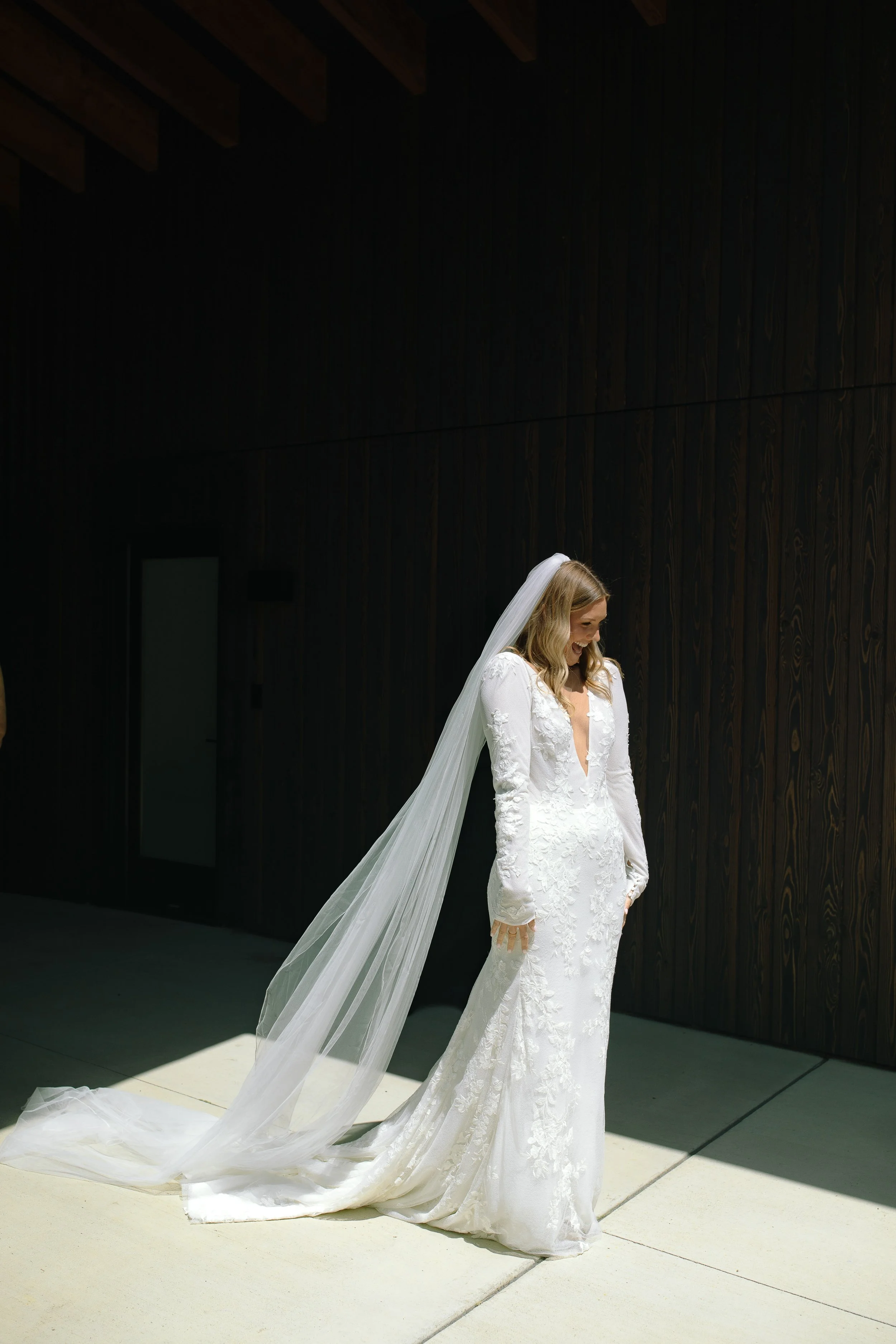 Bride in a lace long-sleeve gown with a long veil, photographed in bright sunlight against the dark wood exterior at Jorgensen Farm The Gardens in Columbus, Ohio.