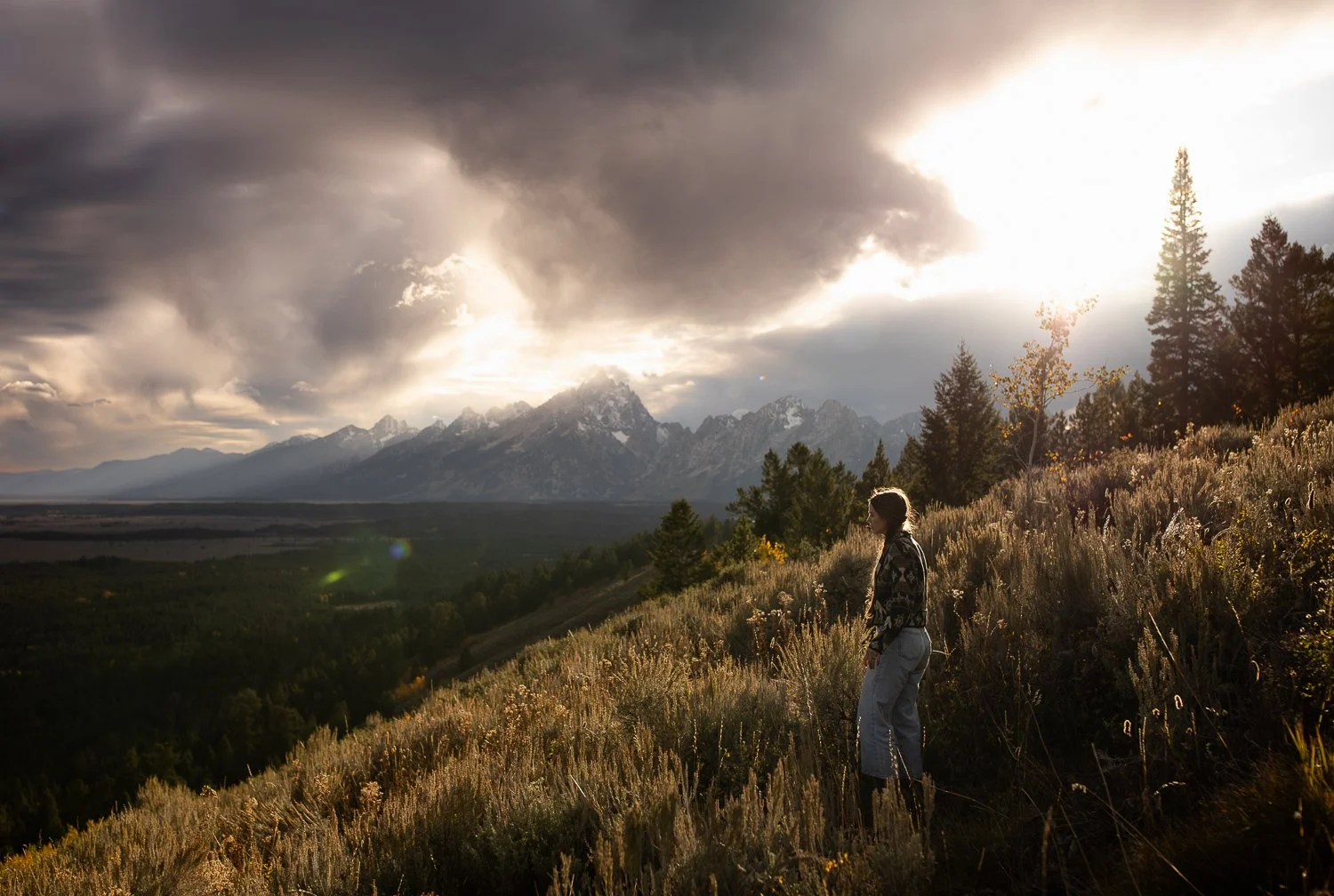 Person standing on a hillside overlook at sunset with dramatic clouds over Grand Teton National Park