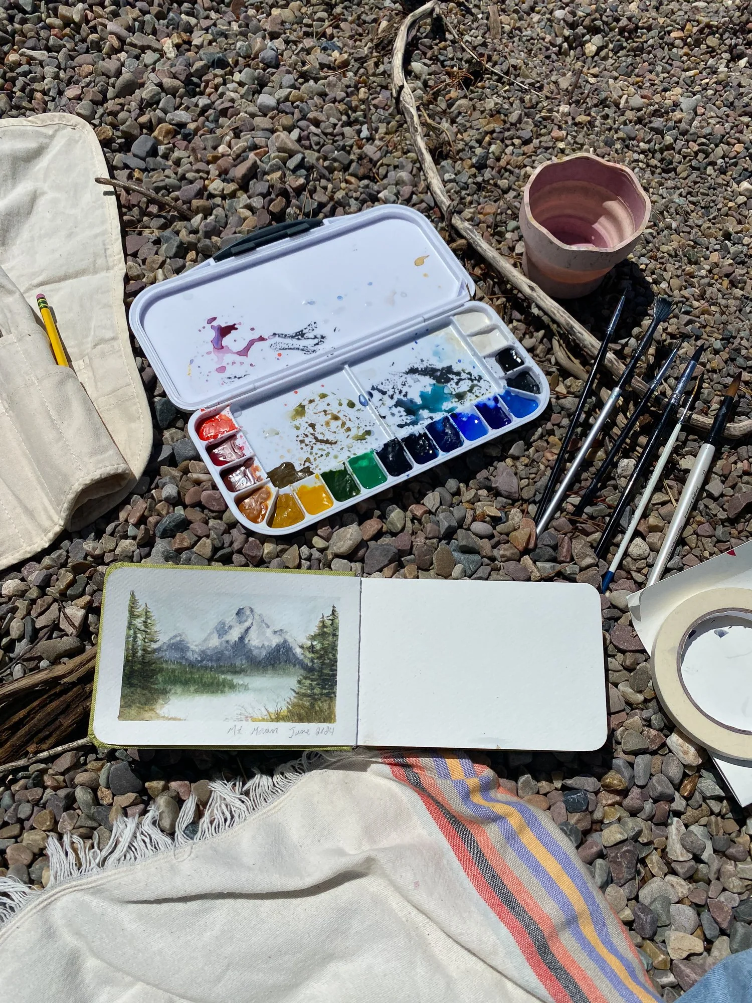 Watercolor painting supplies and sketchbook beside a small Grand Teton landscape painting outdoors
