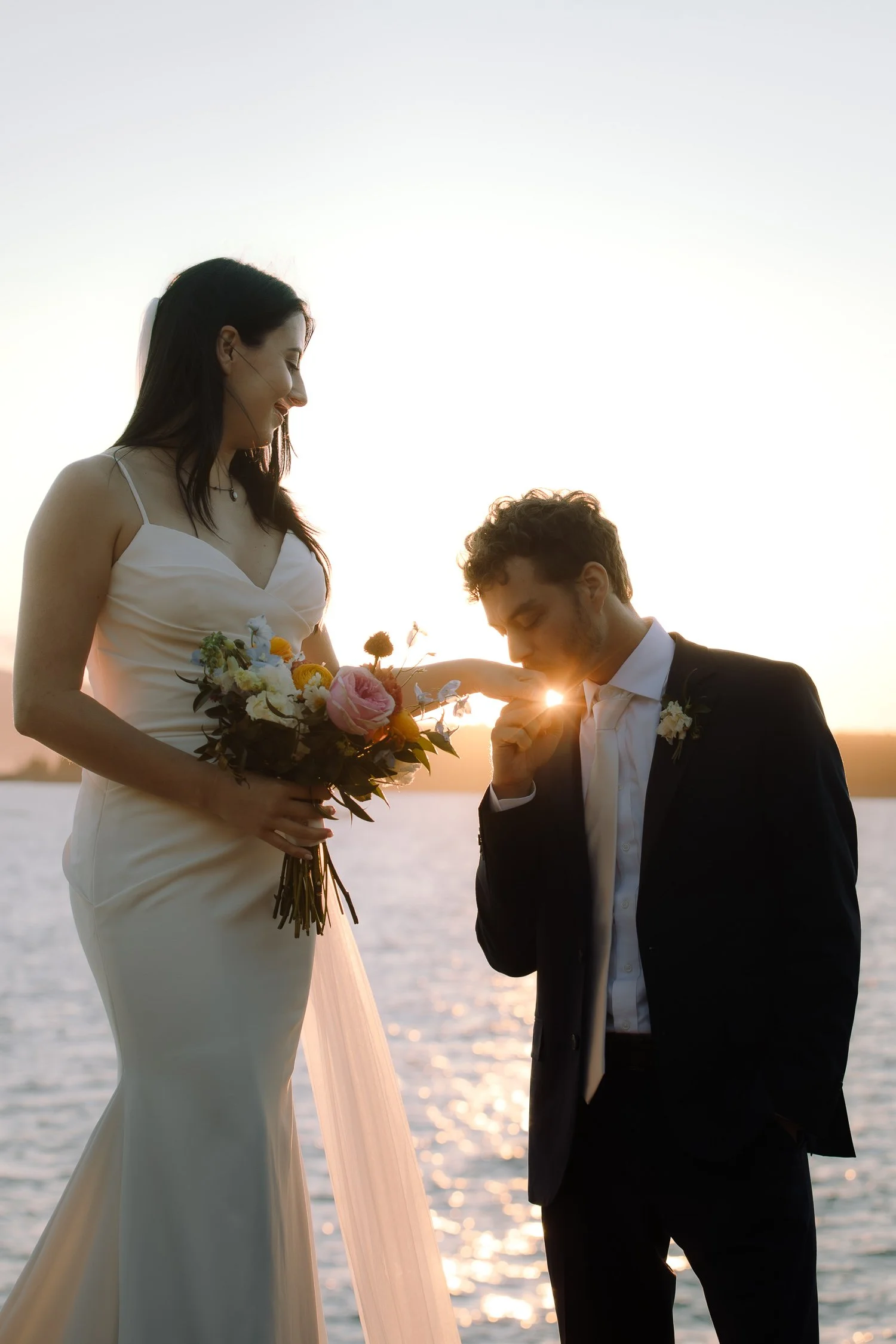 Groom kissing the bride’s hand as warm sunset light reflects off the ocean during an Acadia elopement