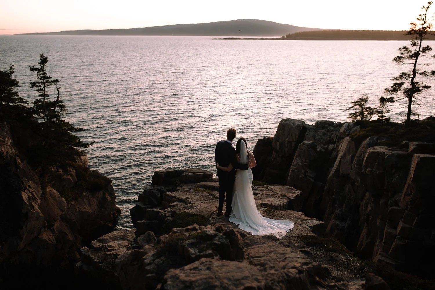 Couple standing together on rocky cliffs overlooking the ocean at sunset during an Acadia elopement