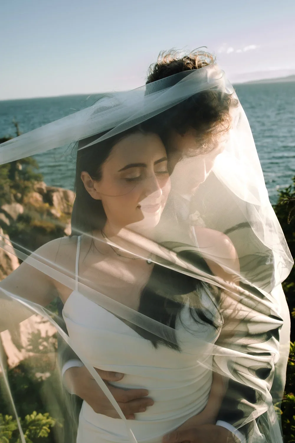 Intimate elopement portrait of a couple wrapped in a veil along the rocky coast of Acadia National Park