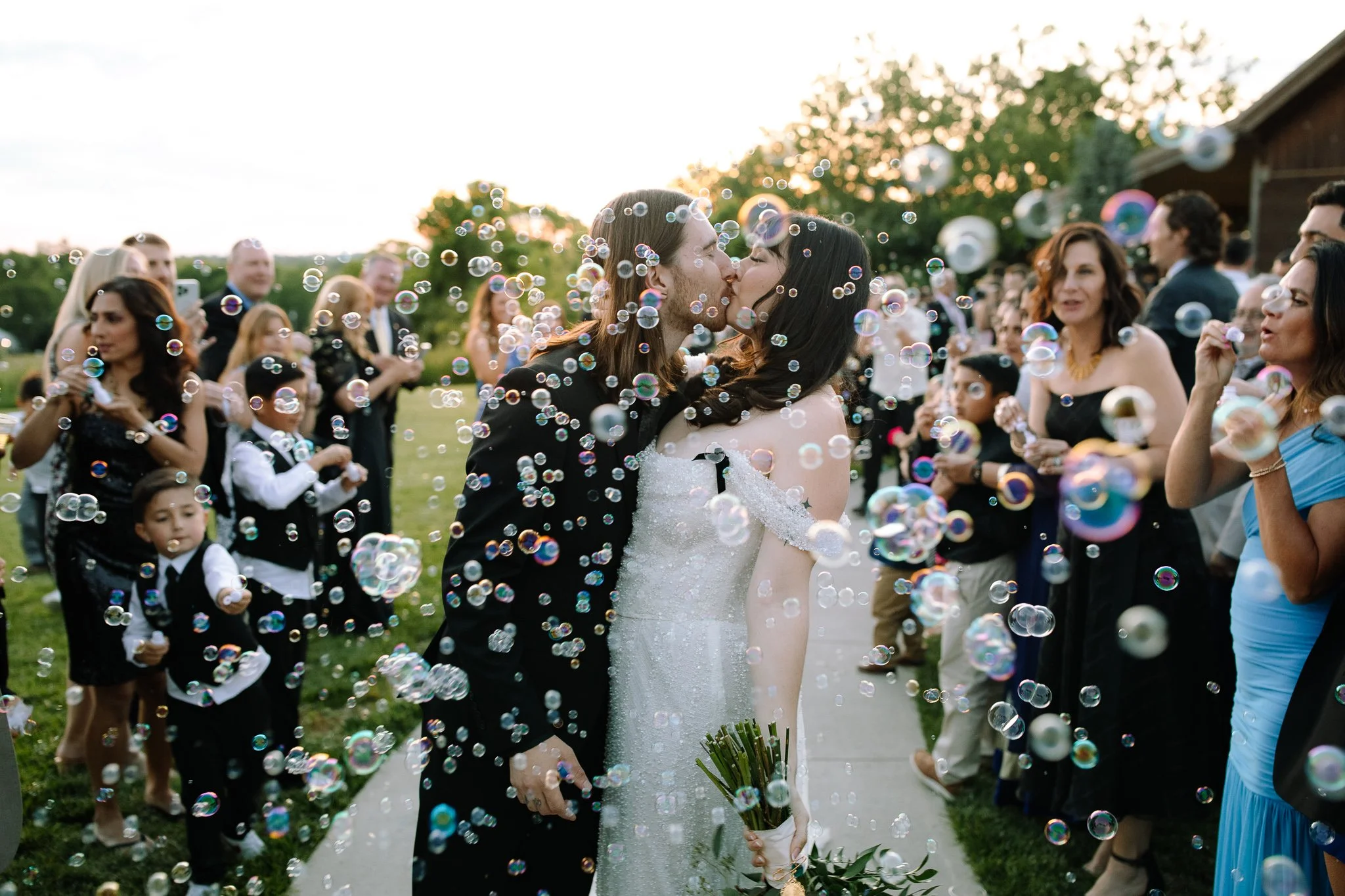Bride and groom share a kiss during a joyful bubble exit at Stone Valley Meadows near Dayton, Ohio, surrounded by guests on an open lawn.