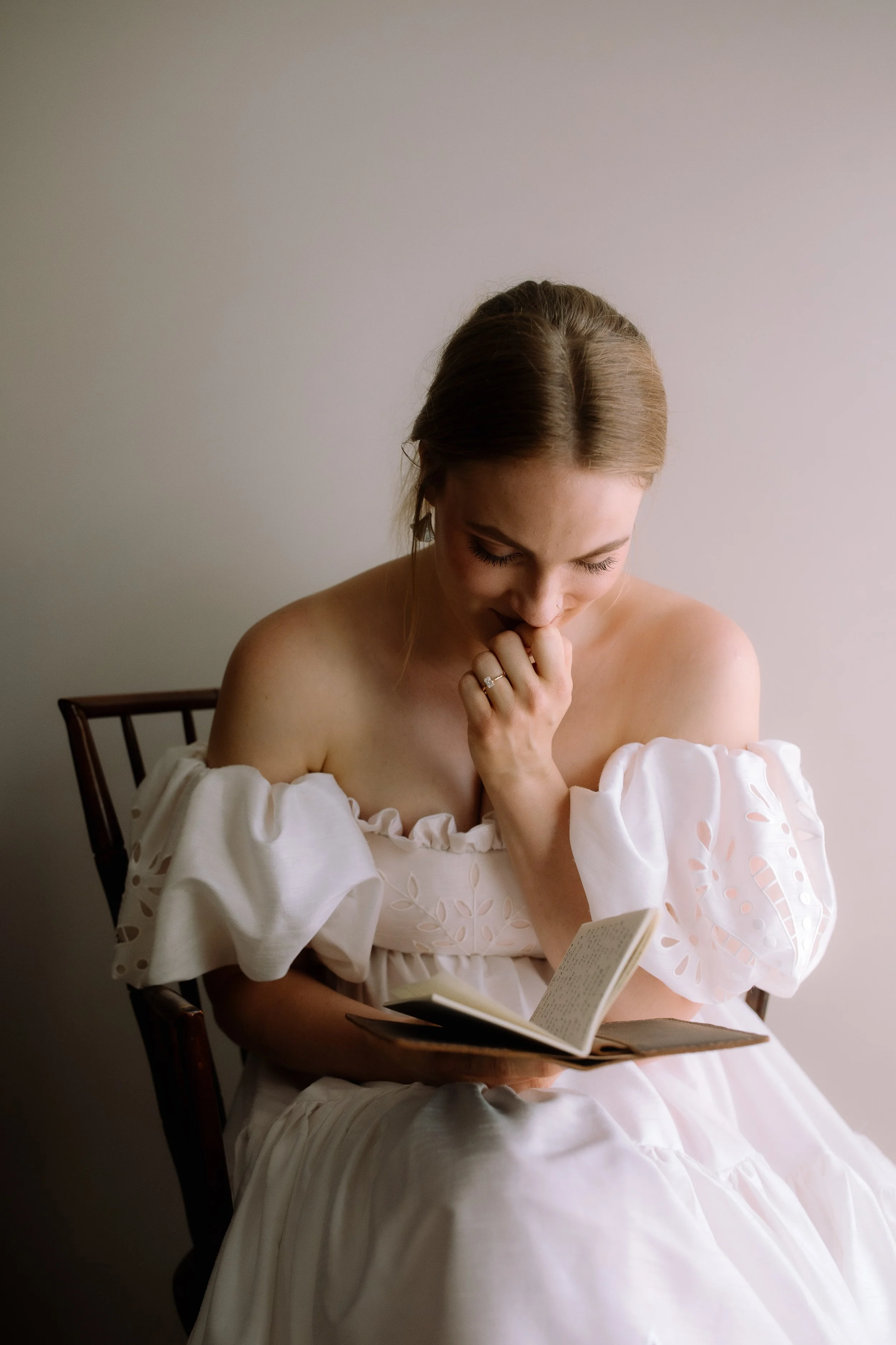 Bride sitting in soft window light reading and writing in her vow book on the morning of her wedding.