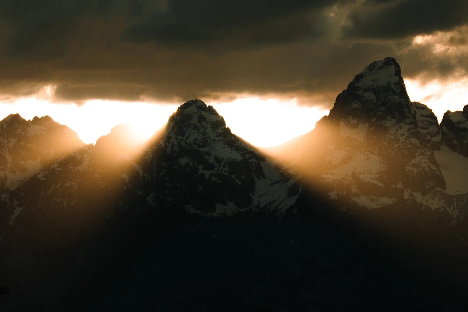Sun breaking through clouds and illuminating the peaks of the Grand Teton mountain range at sunset.