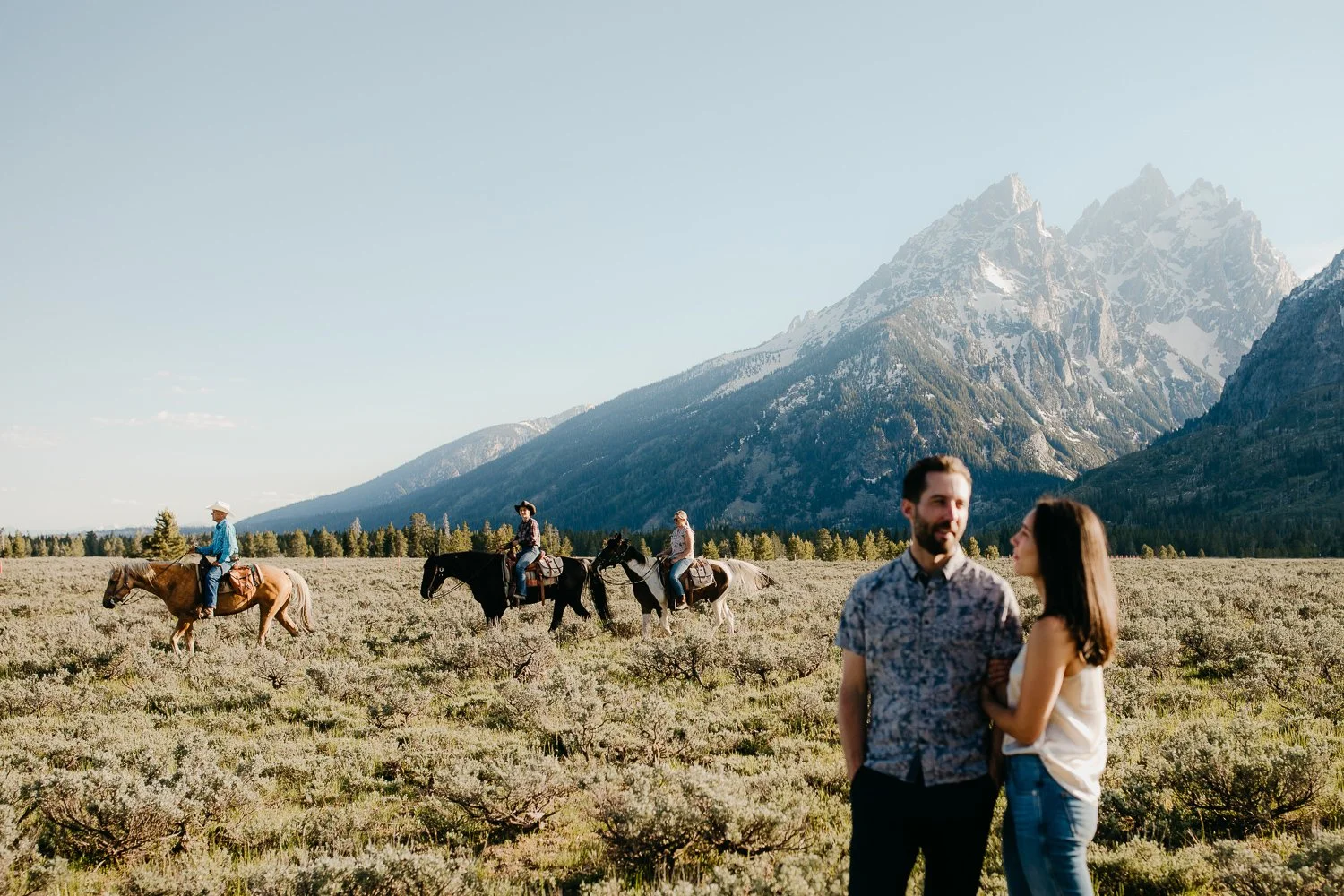 Couple walking together during engagement session with horses in the distance in Jackson Hole